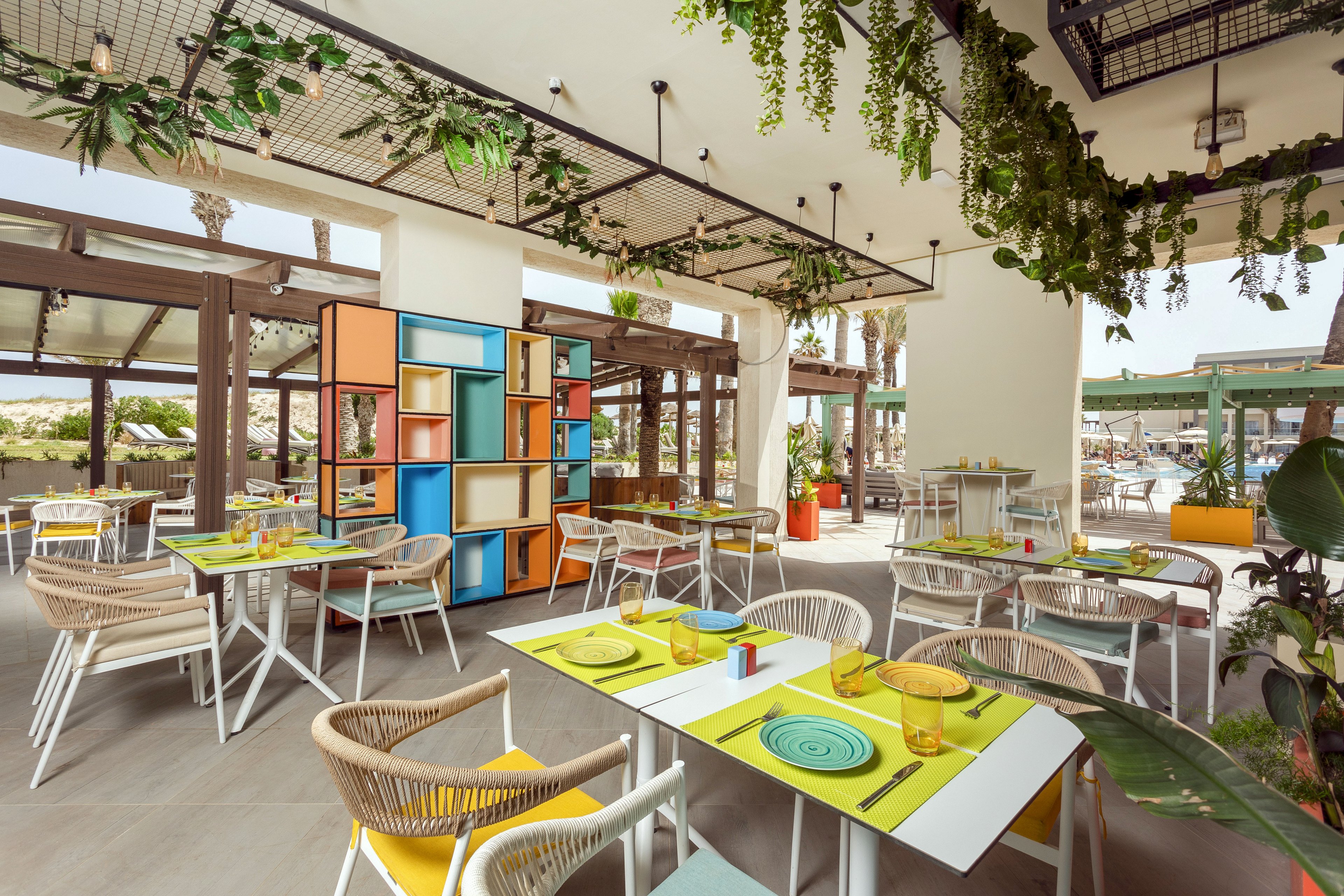 An open-air restaurant features neatly arranged tables with bright yellow placemats and modern wicker chairs. Colorful shelving units and hanging greenery add a playful, contemporary touch to the space. Large windows and a partially covered terrace allow natural light to fill the dining area. The relaxed setting suggests a casual meal near a beach or resort environment.
