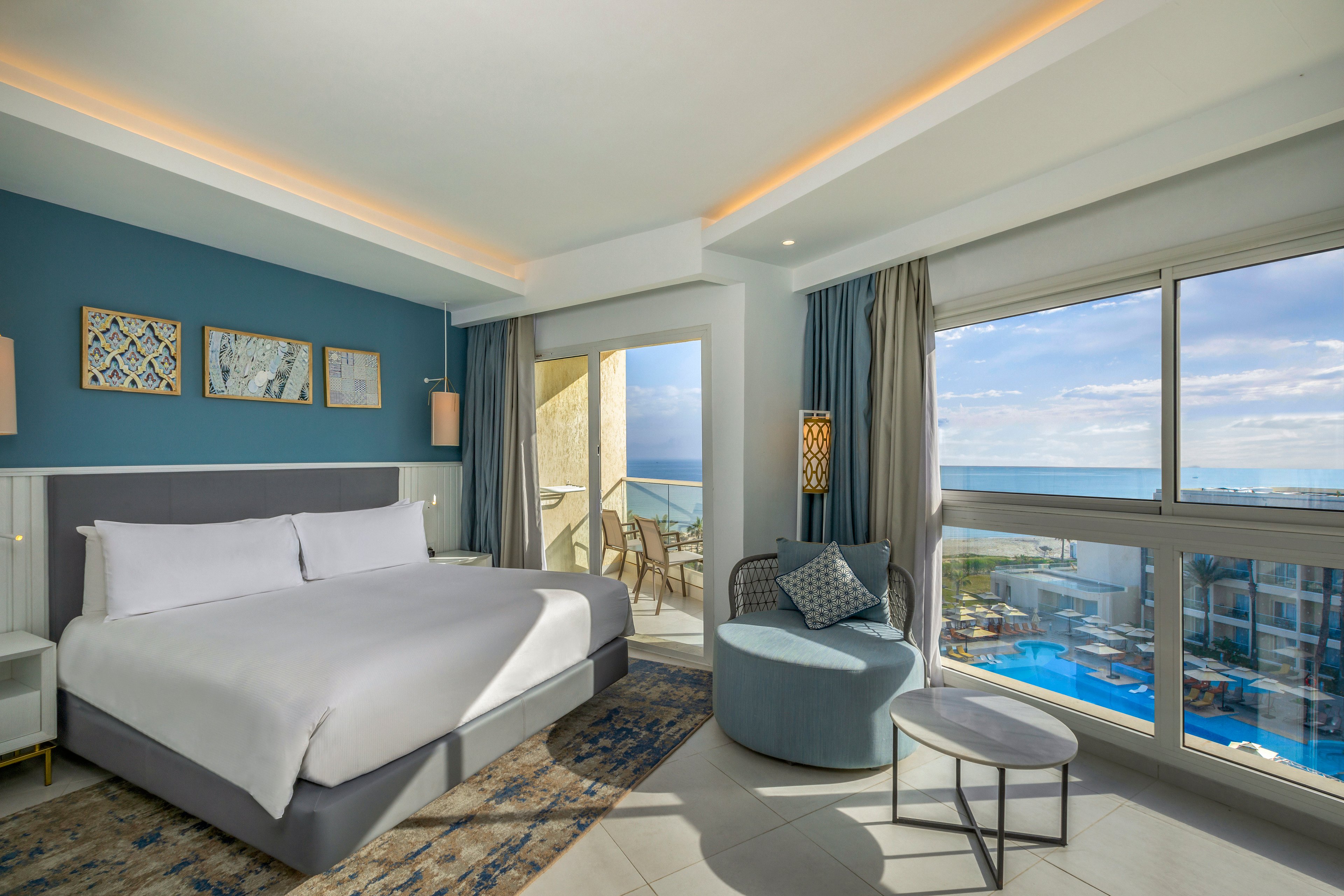Bright contemporary hotel room features a large bed with crisp white linens and blue accent wall art. A cozy seating area with an upholstered chair and small tables sits near wide windows. Floor-to-ceiling glass doors open to a balcony overlooking a swimming pool and expansive ocean views. Soft natural light and neutral tones create a calm, relaxing atmosphere ideal for a beachside stay.