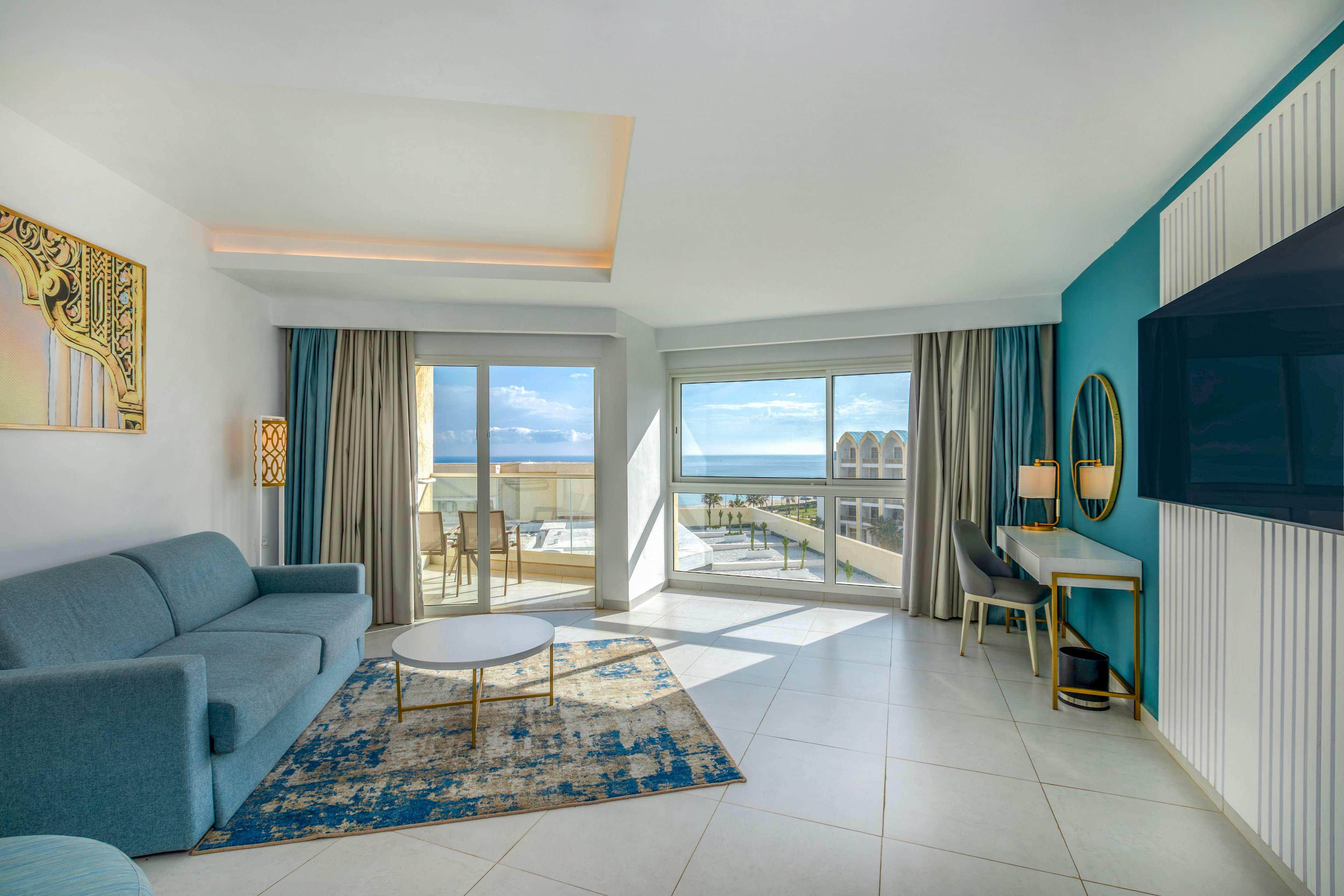 Spacious modern hotel suite features a blue sofa, round coffee table, and large wall-mounted television. Floor-to-ceiling windows and a glass door open to a balcony overlooking the ocean and nearby buildings. A patterned rug, teal accent wall, and contemporary artwork add color and style to the neutral tiled floor. A small desk area with chairs and lamps completes the airy, vacation-ready interior.