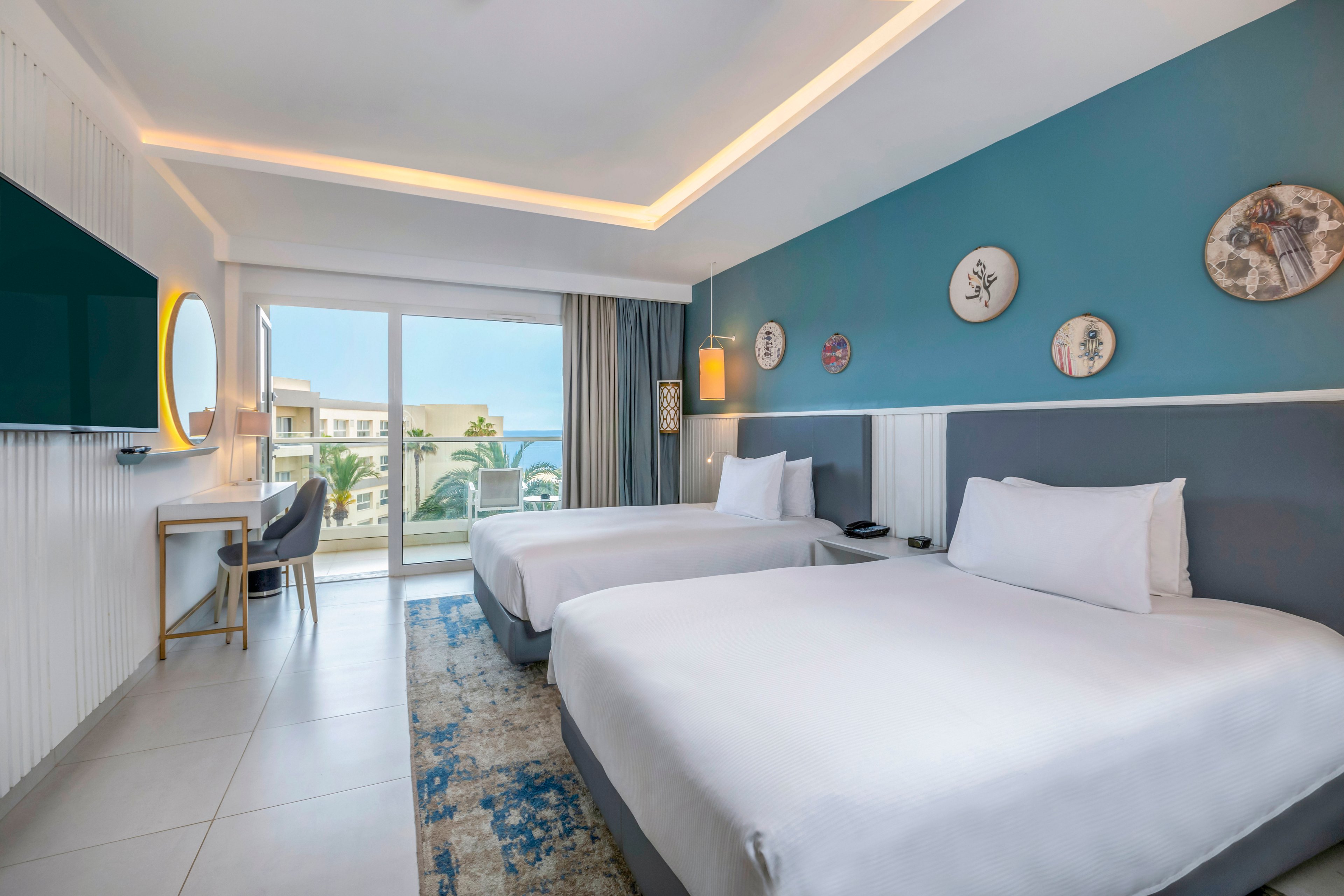 Bright contemporary hotel room featuring two neatly made twin beds with white linens. A blue accent wall with decorative plates and warm bedside lighting adds a stylish touch. Large sliding glass doors open to a balcony with views of palm trees and nearby buildings. A desk area with chair and wall-mounted television complete the comfortable vacation setting.