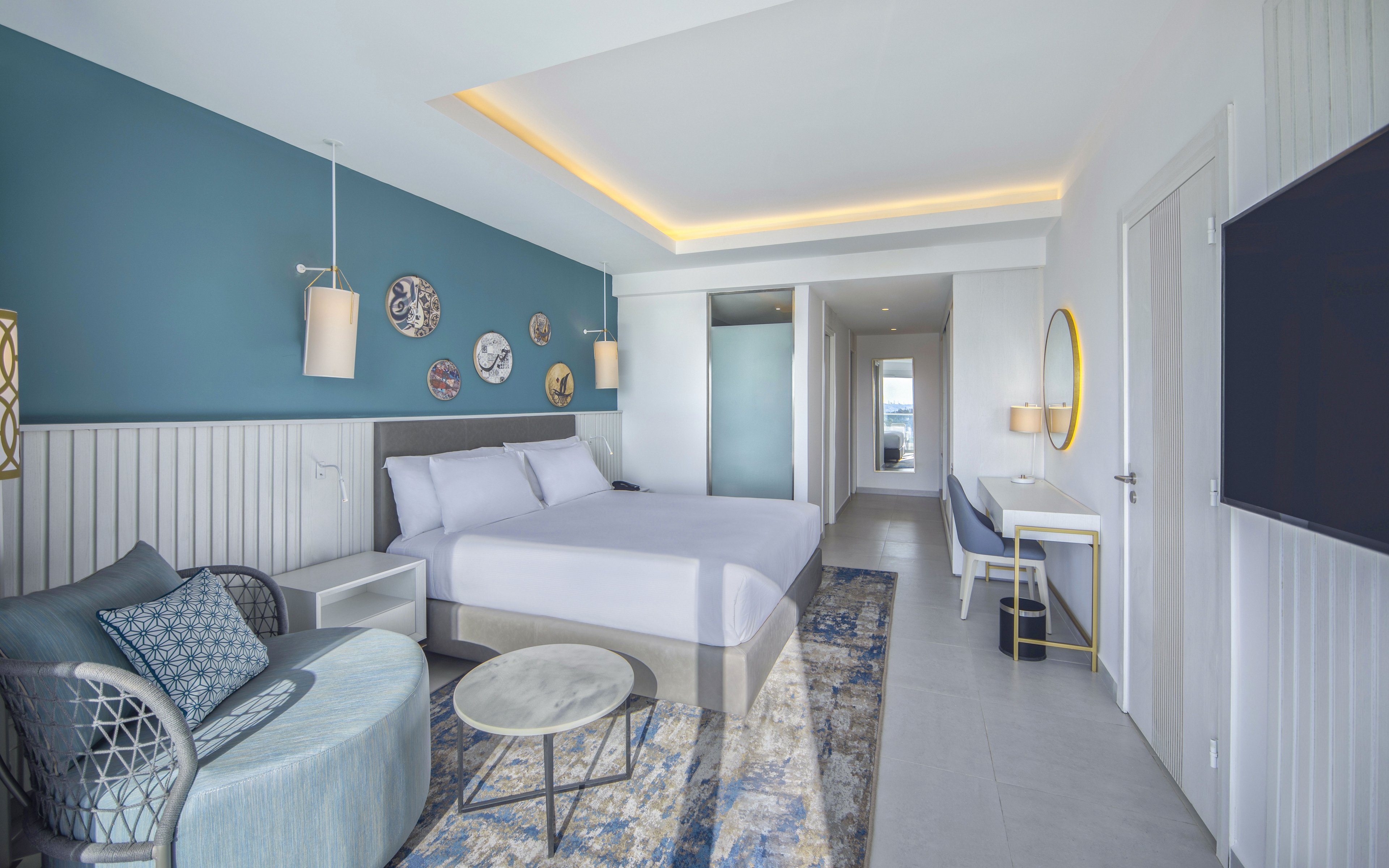 A contemporary hotel room features a large bed with white linens against a teal accent wall. The space includes a cozy seating area with a round ottoman, side table, and patterned rug. Wall-mounted lamps and decorative plates add visual interest, while a hallway leads to the entrance and bathroom area. Soft indirect ceiling lighting and a large flat-screen TV create a bright yet relaxing atmosphere.