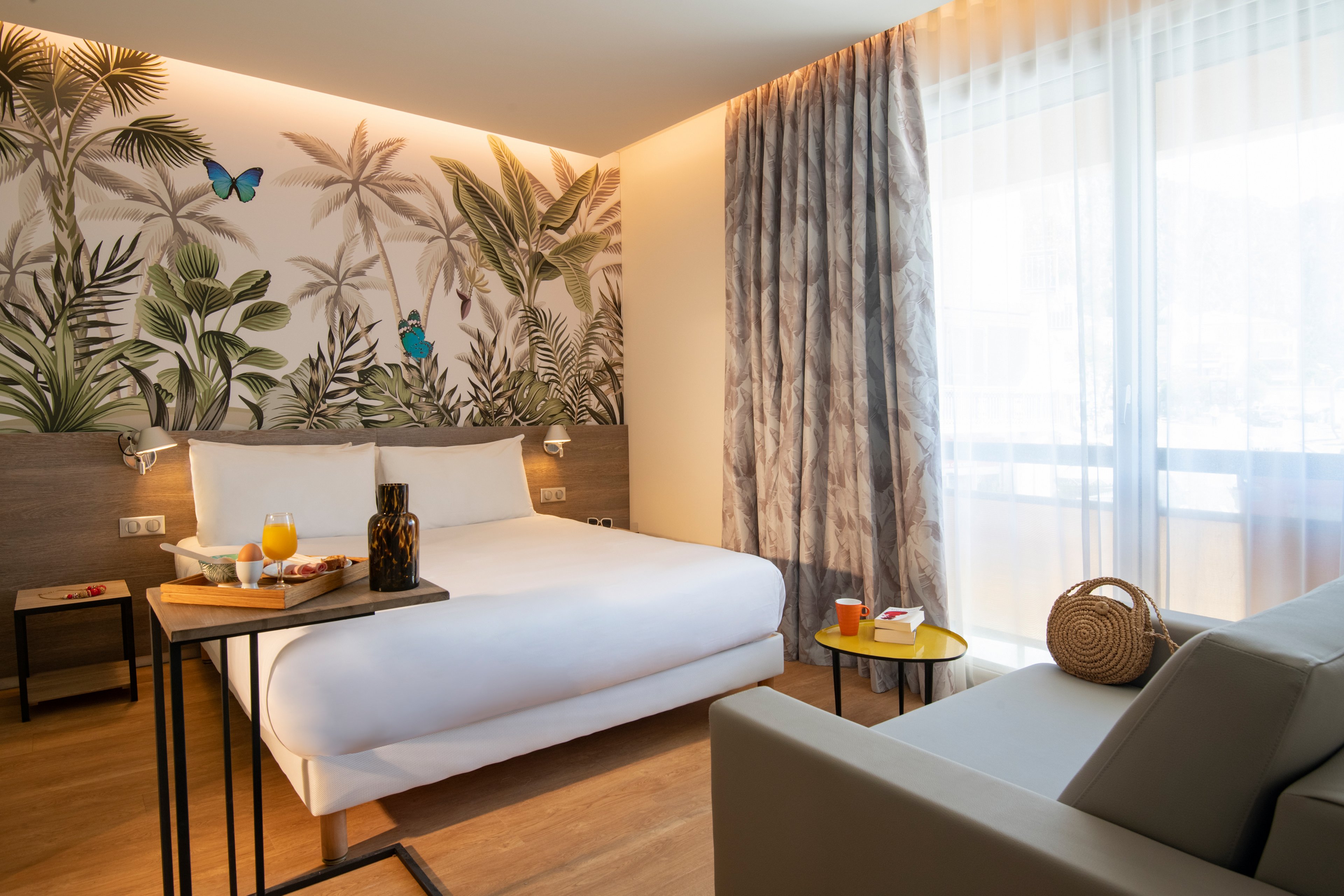 A stylish hotel room featuring a tropical-themed wall mural with lush greenery and butterflies. The setting includes a neatly made bed, a cozy seating area, and a table with breakfast items. Natural light streams through sheer curtains, creating a warm and inviting atmosphere.