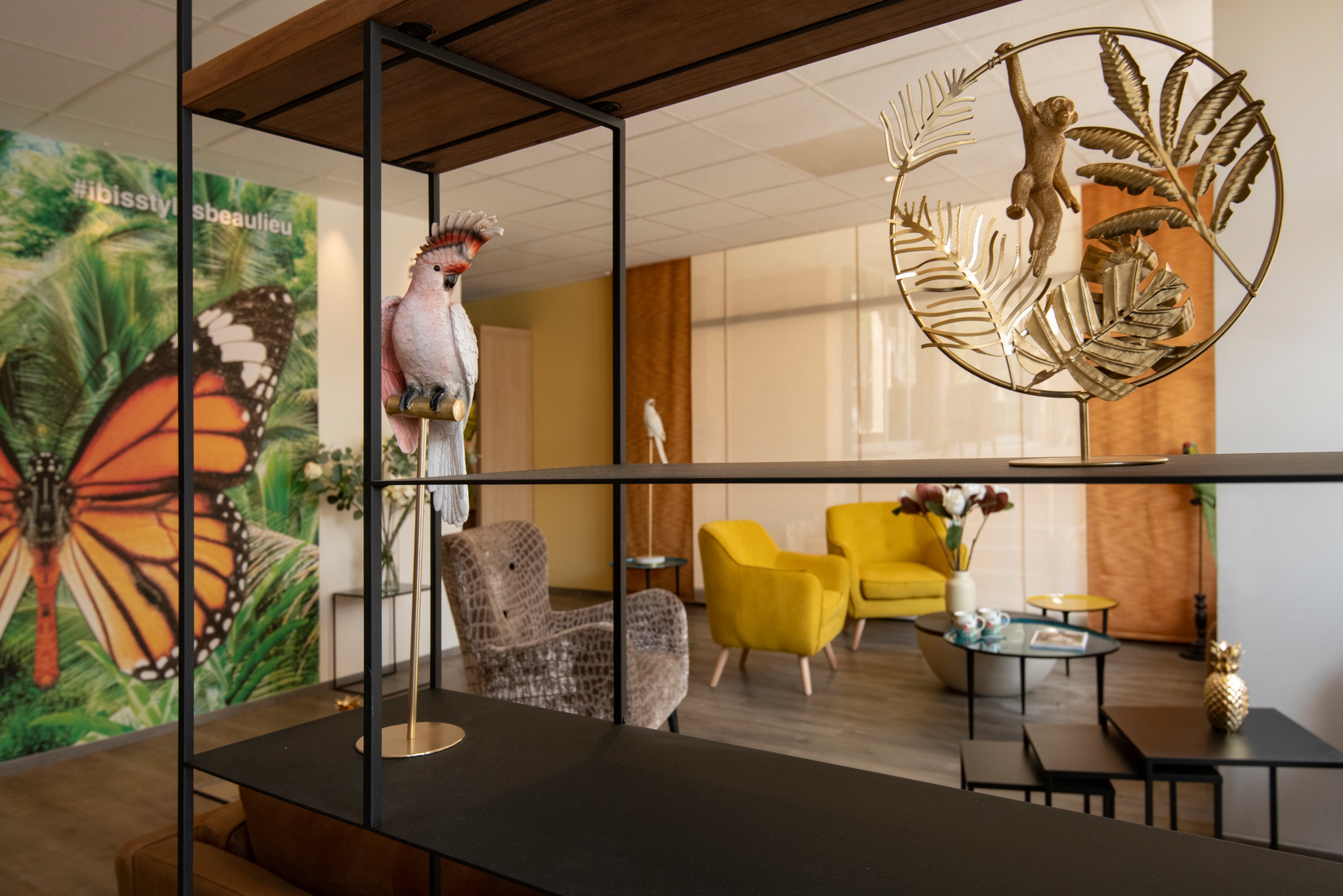A contemporary lounge area featuring vibrant yellow chairs and a decorative parrot sculpture. The setting includes tropical-themed visuals, such as a butterfly mural with visible text 'Ibissy bejeweled'. The space is accented with metallic and botanical designs, creating a warm and inviting atmosphere.