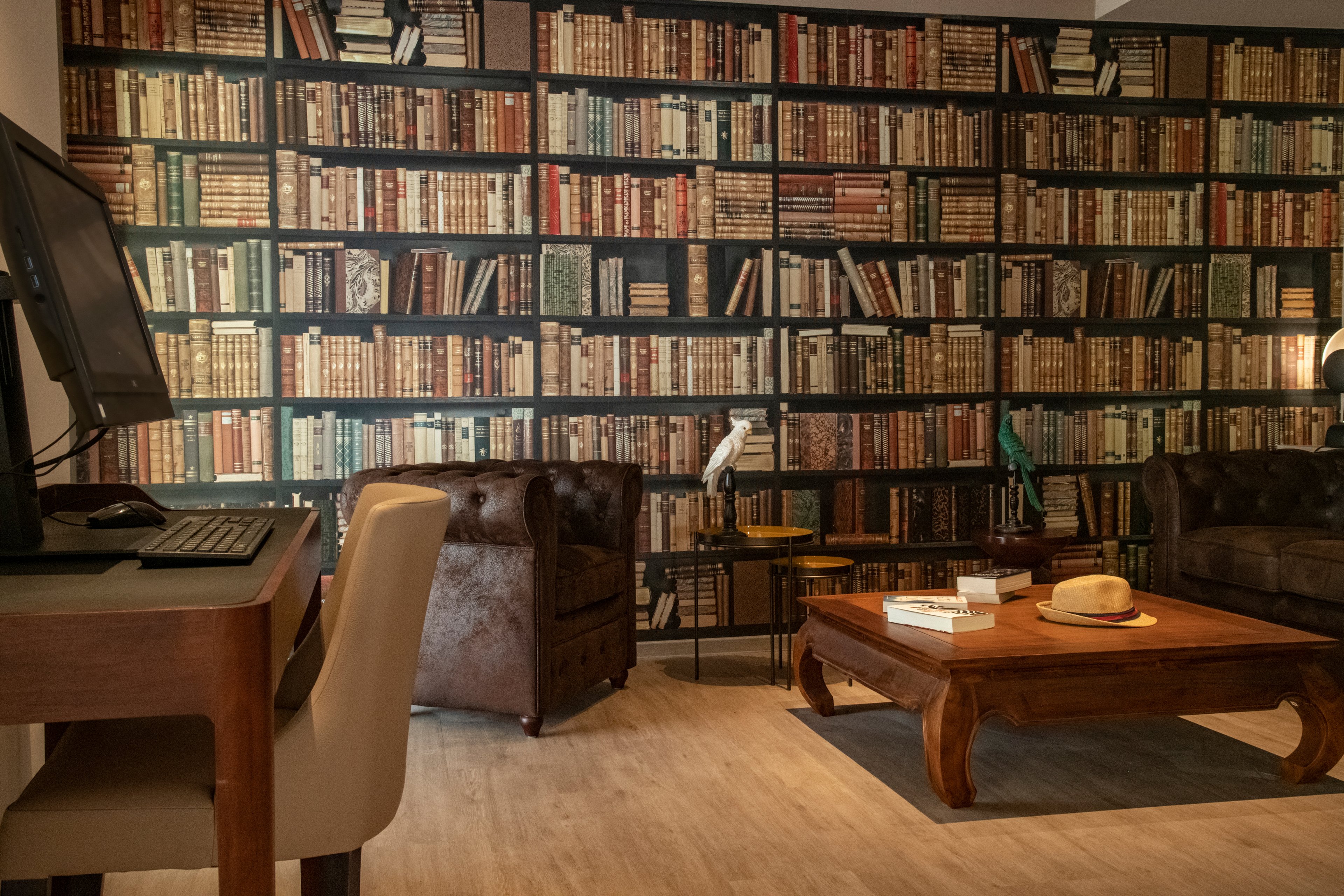 A warm and inviting room featuring a wall covered with a library-themed wallpaper showcasing rows of books. The setting includes leather armchairs, a wooden coffee table, and a desk with a computer. The ambiance is cozy and classic, ideal for reading or working in a relaxed environment.