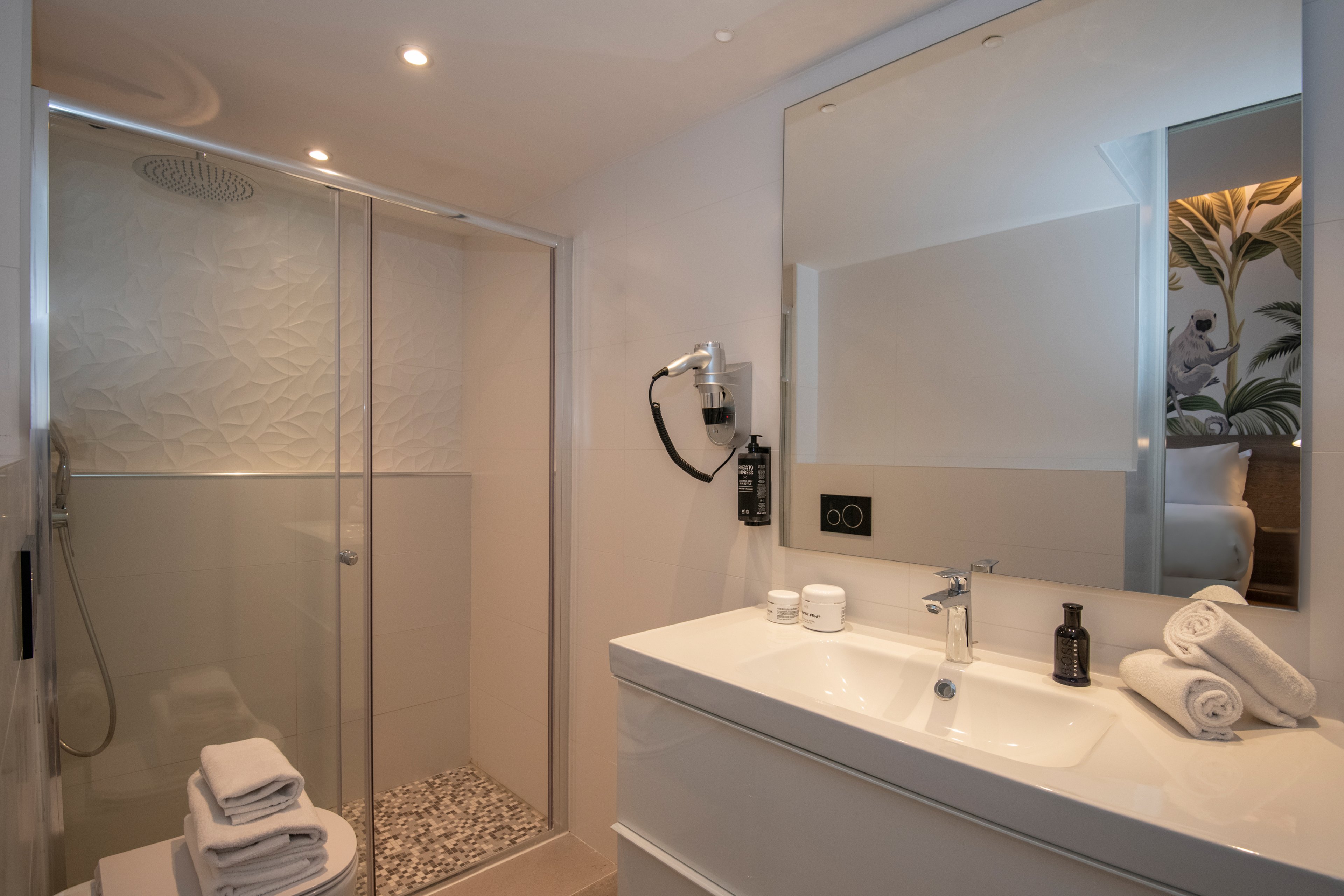 A sleek and contemporary bathroom featuring a glass shower enclosure with a textured wall design. The setting includes a white countertop sink with neatly folded towels and a black soap dispenser. Warm lighting enhances the clean and minimalist aesthetic, with a glimpse of a bedroom visible in the mirror reflection.