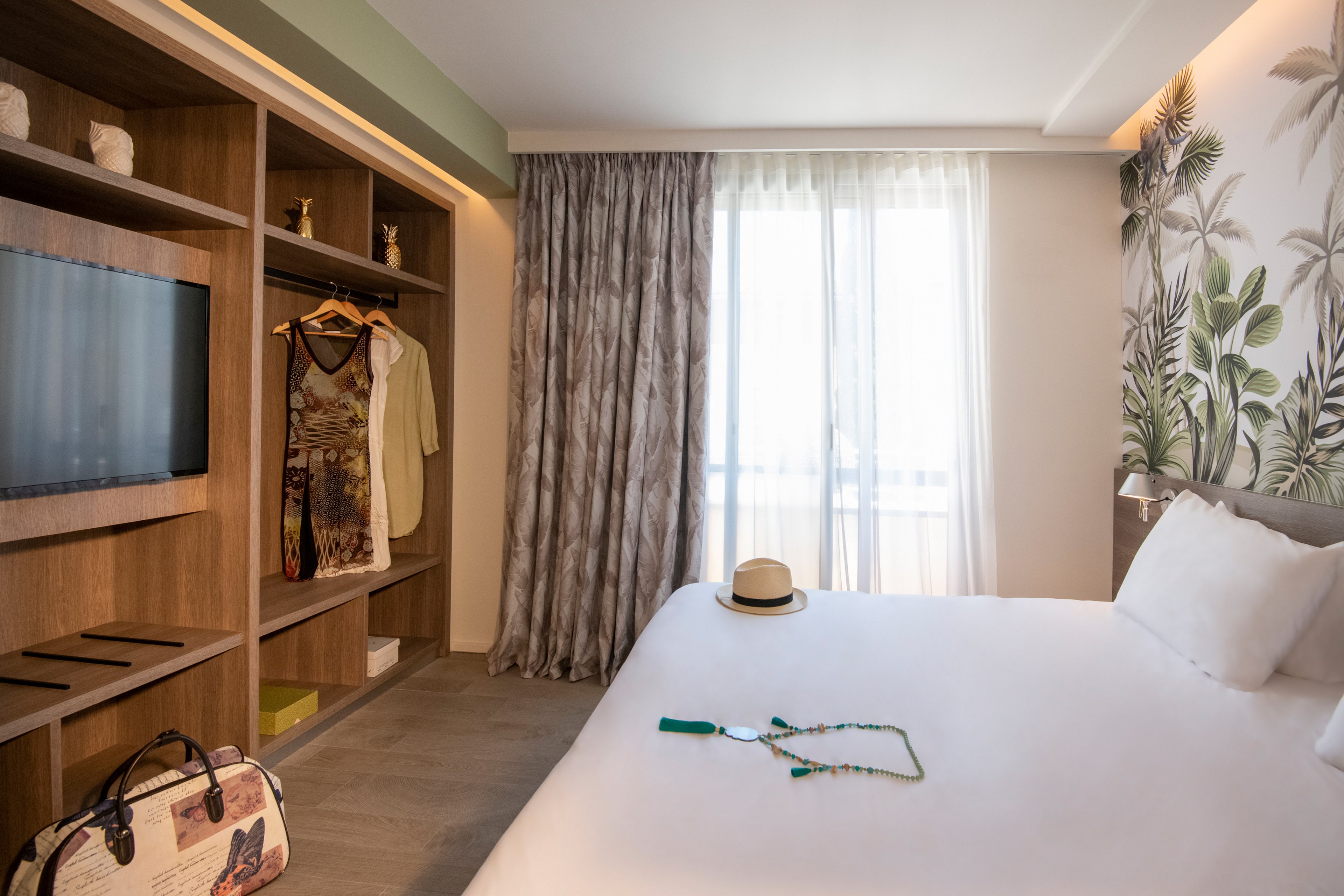 A bright and stylish hotel room featuring tropical-themed wallpaper and wooden furnishings. The setting includes a neatly made bed, open wardrobe with hanging clothes, and a window with sheer curtains allowing natural light. A straw hat, necklace, and travel bag are visible, adding a vacation vibe.