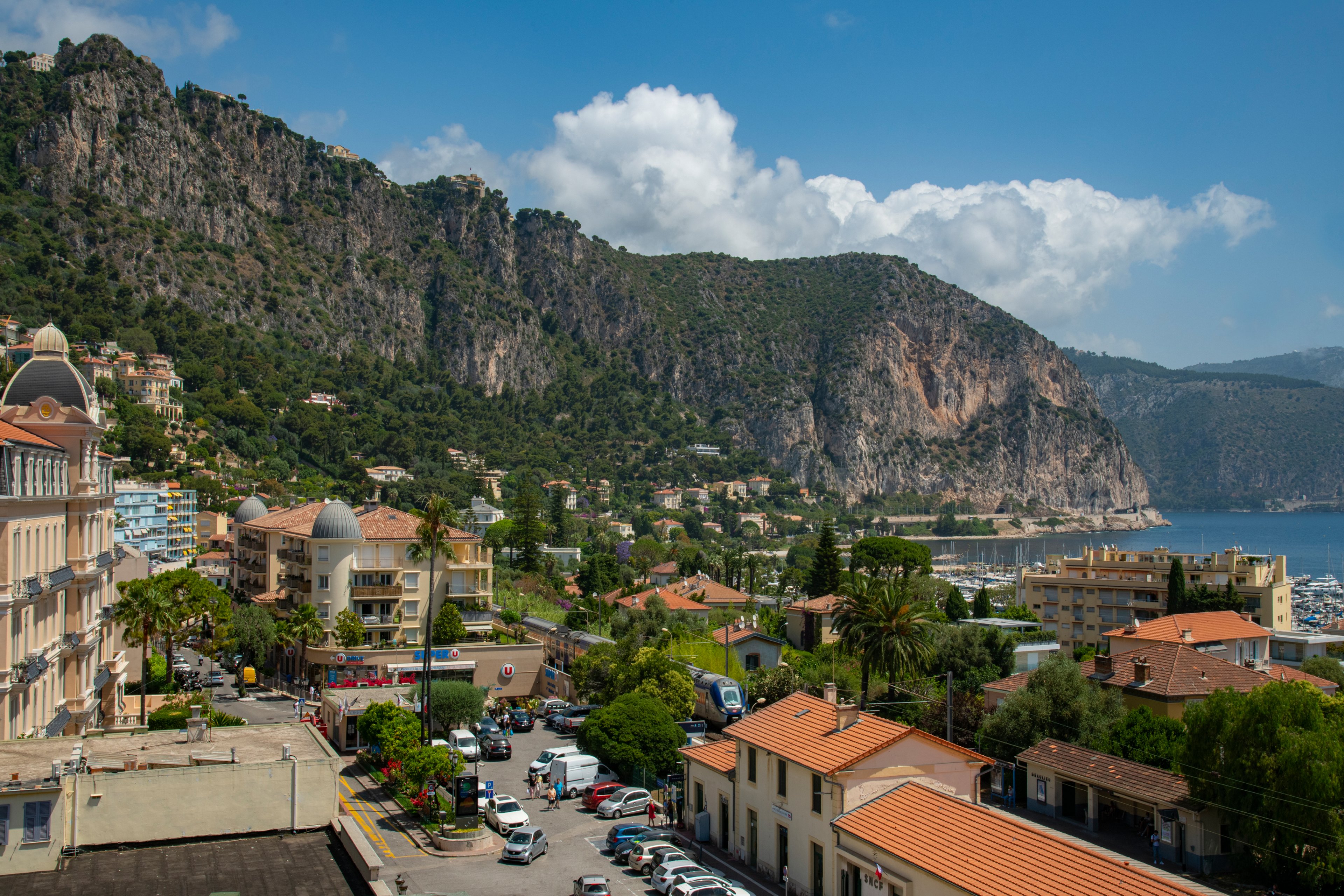 A picturesque coastal town nestled against rugged mountains under a bright blue sky. The setting features residential buildings, lush greenery, and a view of the sea. The image captures a serene atmosphere with clear weather and vibrant colors.