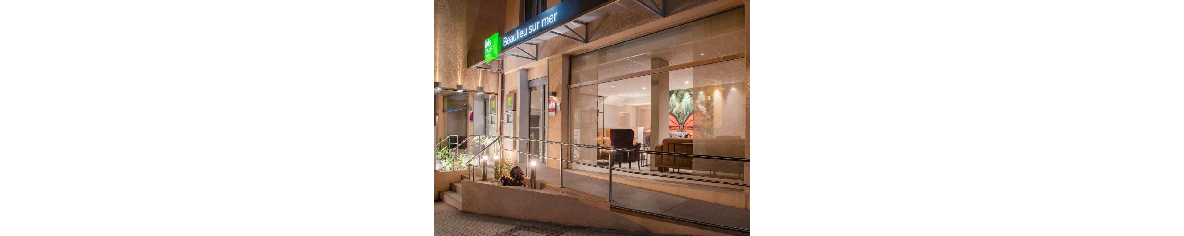A contemporary hotel facade illuminated at night, featuring the text 'Beaulieu sur Mer' clearly visible on signage. The interior showcases a cozy seating area with warm lighting and tropical-themed decor. The setting includes a ramp and steps leading to the entrance, surrounded by minimal landscaping.