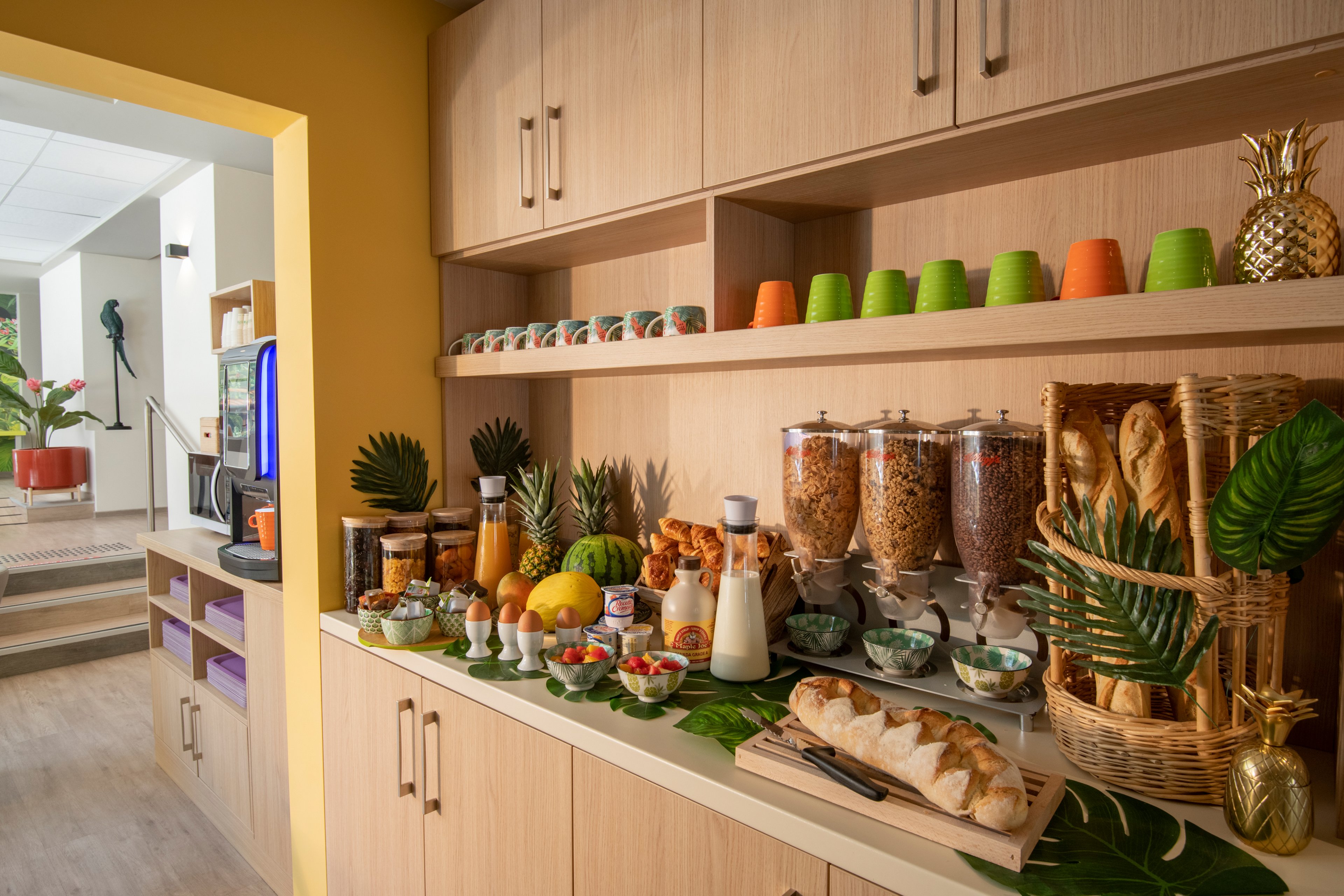 A well-organized breakfast buffet featuring cereals in dispensers, fresh bread, and tropical fruits. The setting is a bright and modern kitchen area with wooden cabinets and colorful cups. Decorative elements include green leaves and a wicker basket, creating a fresh and inviting atmosphere.