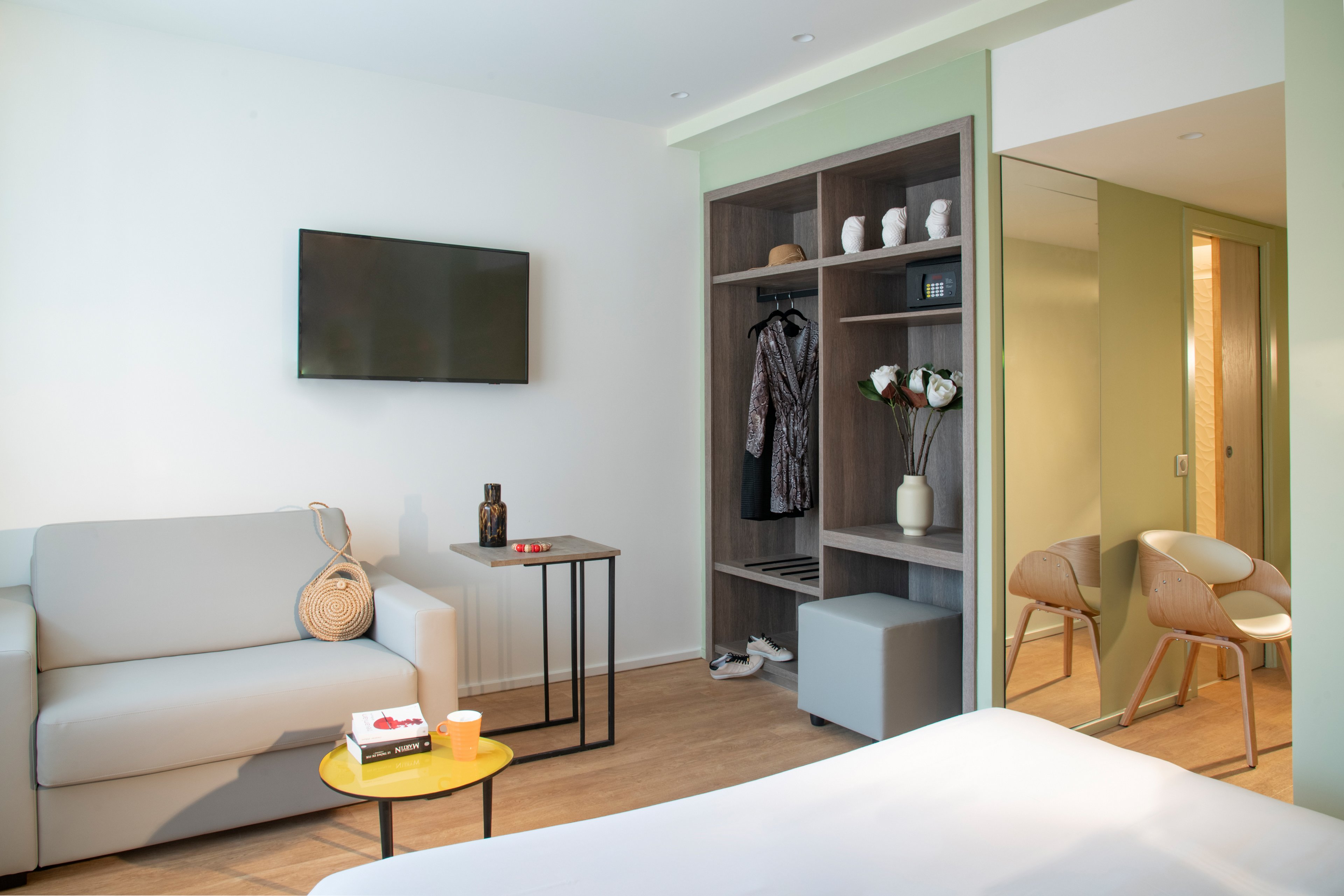 A contemporary hotel room featuring a comfortable sofa, a wall-mounted television, and an open wardrobe with decorative items. The setting includes a small table with a cup and book, creating a relaxed atmosphere. Neutral tones and soft lighting enhance the inviting mood of the space.