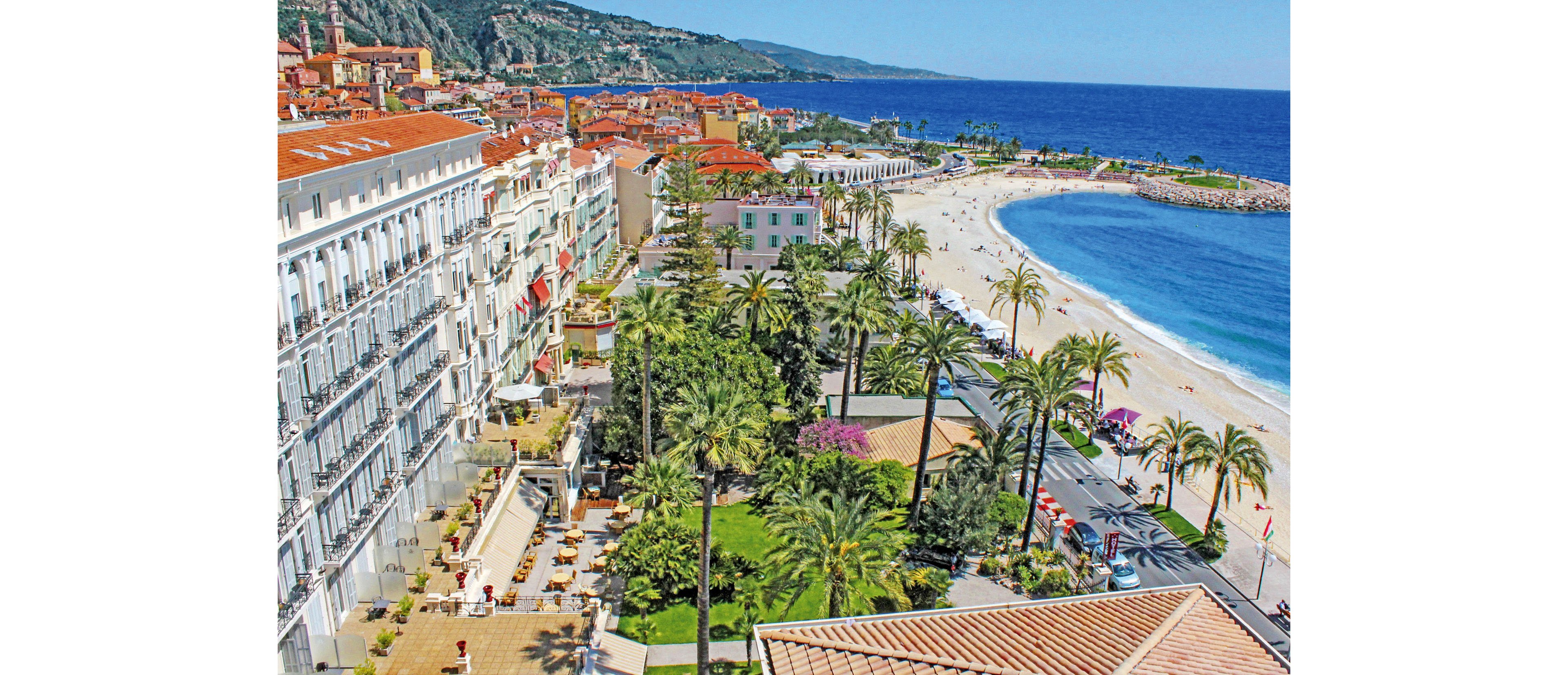 A vibrant coastal cityscape shows a long beachfront lined with palm trees and elegant multi-story buildings. The wide sandy beach curves along the bright blue sea, with people relaxing and walking near the shoreline. A busy promenade and road run parallel to the coast, adding an urban feel to the scenic seaside setting. Hills and colorful rooftops rise in the background, enhancing the picturesque Mediterranean atmosphere.