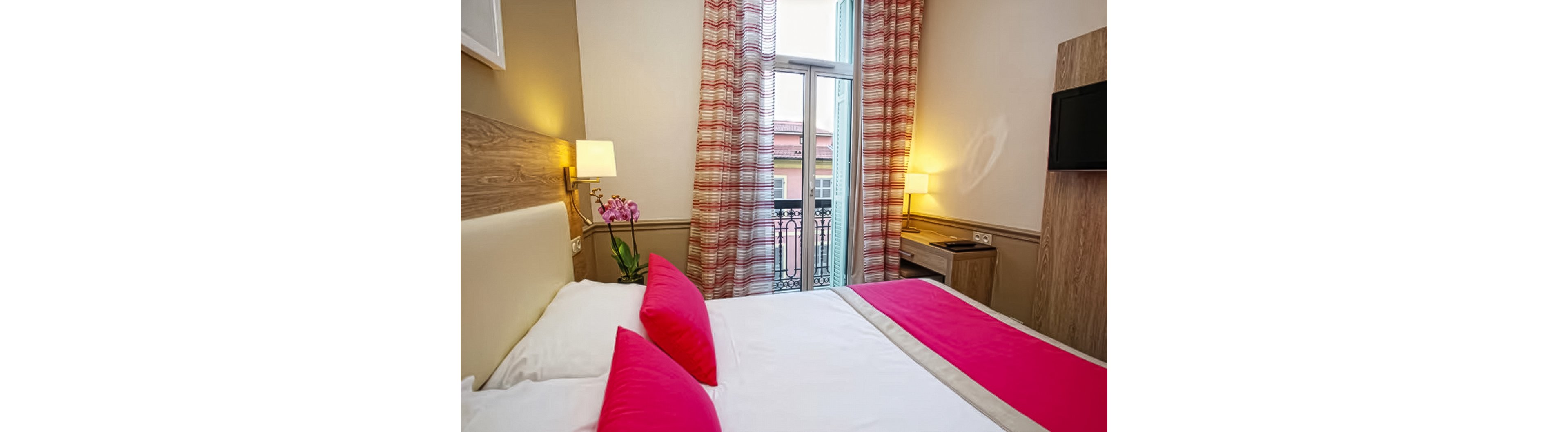 A contemporary hotel room features a neatly made double bed with white linens and vivid pink accents. The space includes wooden furniture, a wall-mounted television, and bedside lamps casting warm light. Patterned curtains frame a tall glass door that opens to a small balcony with decorative metal railing and a view of neighboring buildings. A potted orchid adds a touch of color and elegance to the interior decor.