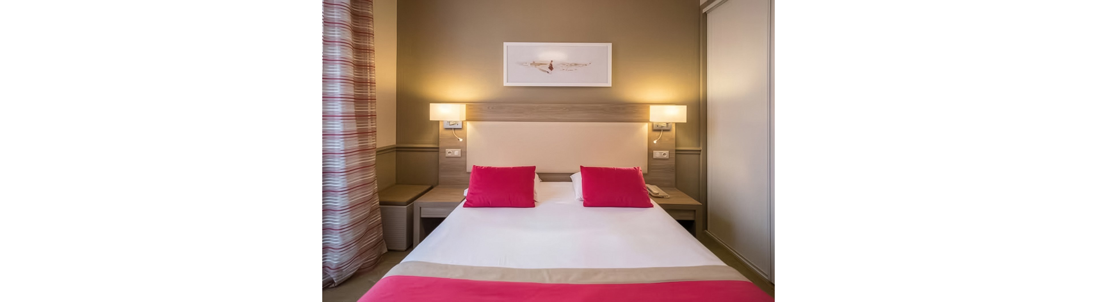 A neatly made double bed with white linens and bright pink pillows is centered in a modern hotel room. Warm beige walls and matching headboard are flanked by two wall-mounted lamps casting soft light. A striped curtain hangs to the left, while bedside tables and a telephone sit on either side of the bed. A minimalist artwork above the headboard completes the calm, contemporary decor.