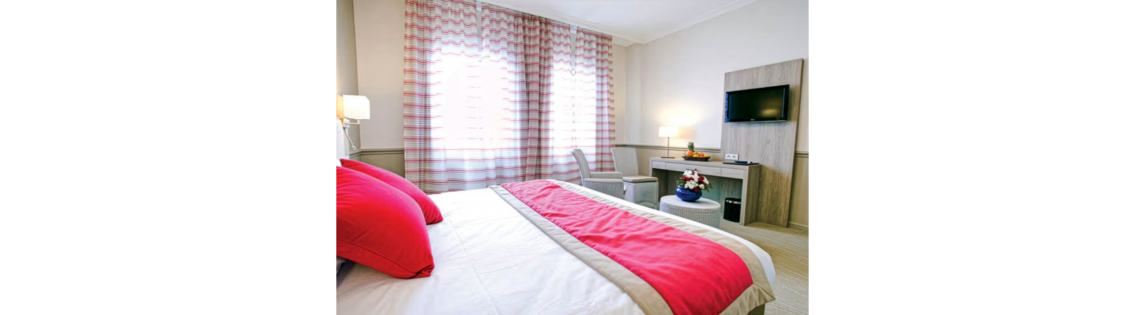 A neatly made bed with white linens and bold red pillows dominates a modern hotel room. Soft natural light filters through patterned curtains covering large windows. A compact desk area with a flat-screen TV, chair, and decorative flower arrangement sits against the wall. The overall decor is contemporary and minimal, featuring neutral tones highlighted by vivid red textiles.