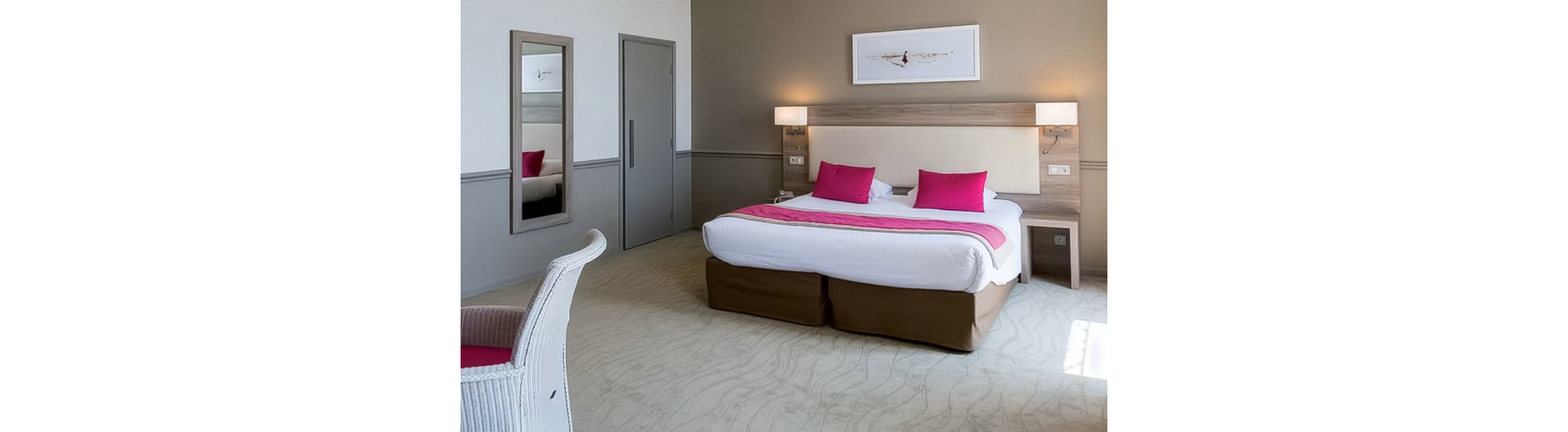 Contemporary hotel room featuring a large double bed with white linens and bright pink pillows and runner. The space includes a light wood headboard with built-in wall lamps and bedside tables. A full-length mirror, simple artwork above the bed, and neutral walls create a clean, minimalist look. Natural light enters from the right, highlighting the patterned carpet and white chair in the foreground.
