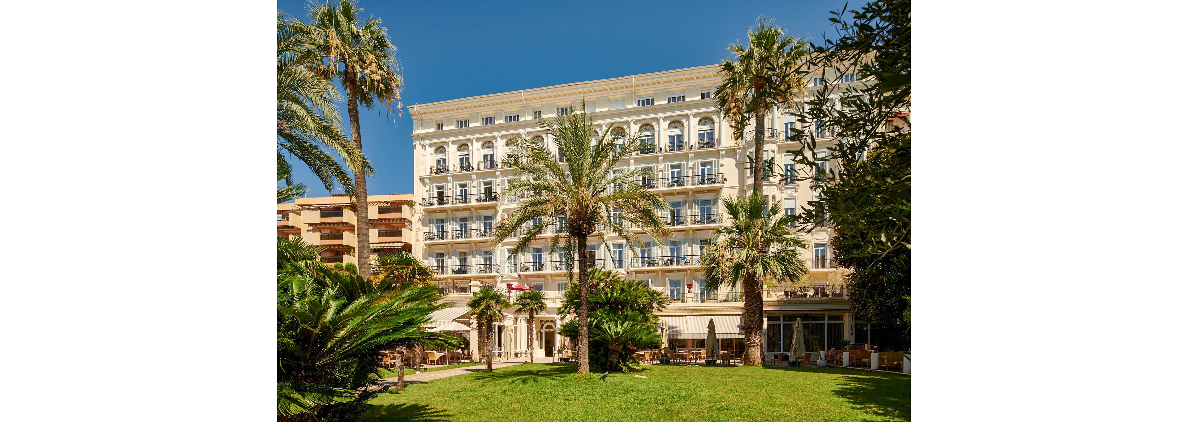 A large multi-story hotel with ornate balconies faces a green garden filled with palm trees and manicured lawns. Outdoor seating and awnings line the ground floor, suggesting a terrace or café area for guests. The building’s light-colored façade contrasts with the bright blue sky, creating a sunny, Mediterranean atmosphere. Adjacent structures and dense foliage frame the central hotel, emphasizing its elegant resort setting.