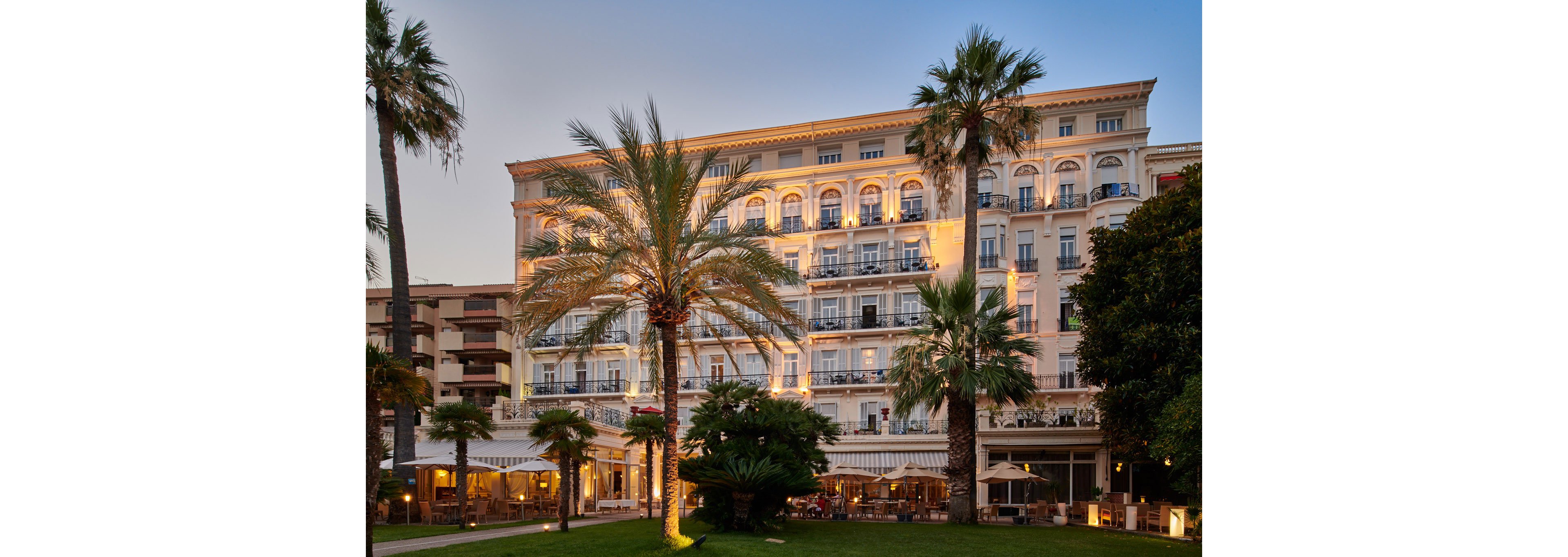 A grand multi-story hotel building is illuminated warmly at dusk, with rows of balconies and arched windows facing a landscaped garden. Tall palm trees and manicured greenery frame the facade, adding a Mediterranean or coastal resort feel. Outdoor seating and canopied terrace areas suggest a restaurant or lounge space on the ground level. The soft evening sky and glowing lights create a relaxed, upscale vacation atmosphere.