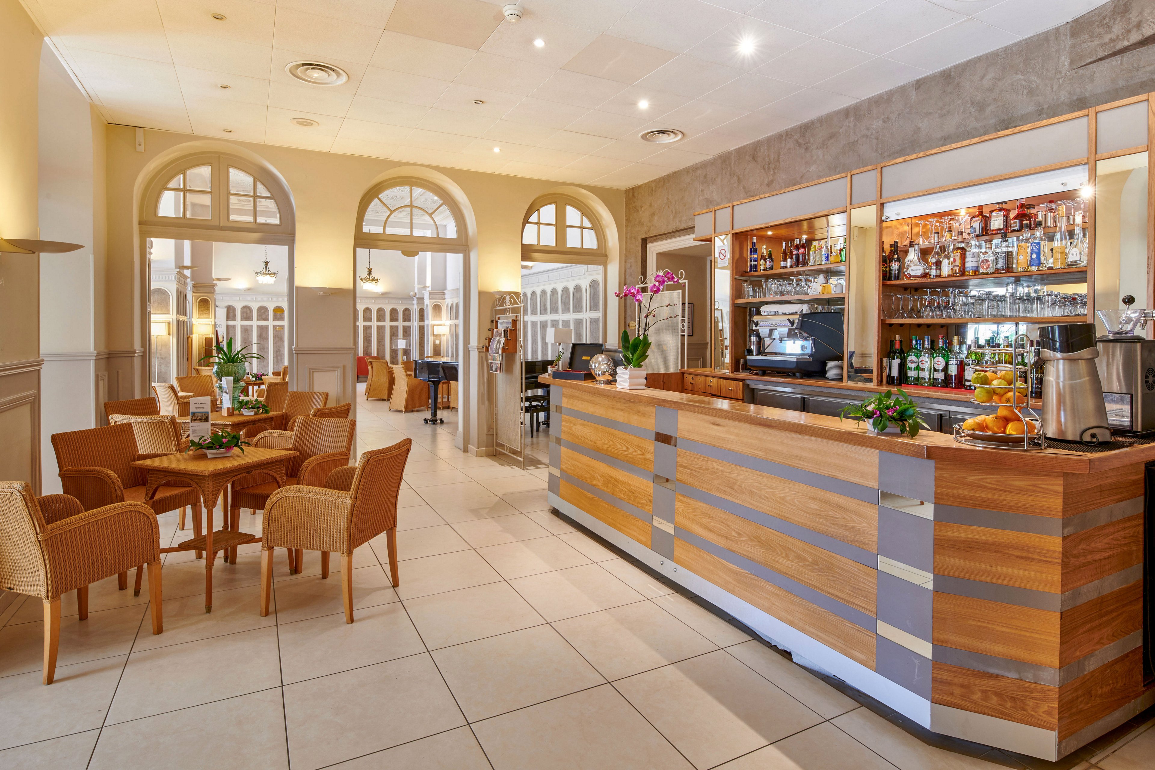 A contemporary hotel bar features a long wooden counter with metal accents and a mirrored back wall filled with bottles and glassware. The space opens into a lounge area with round tables, wicker-style chairs, and decorative plants. Large arched windows and doors allow natural light to brighten the neutral-toned room. The overall atmosphere is welcoming and relaxed, suitable for casual drinks or coffee in a stylish setting.