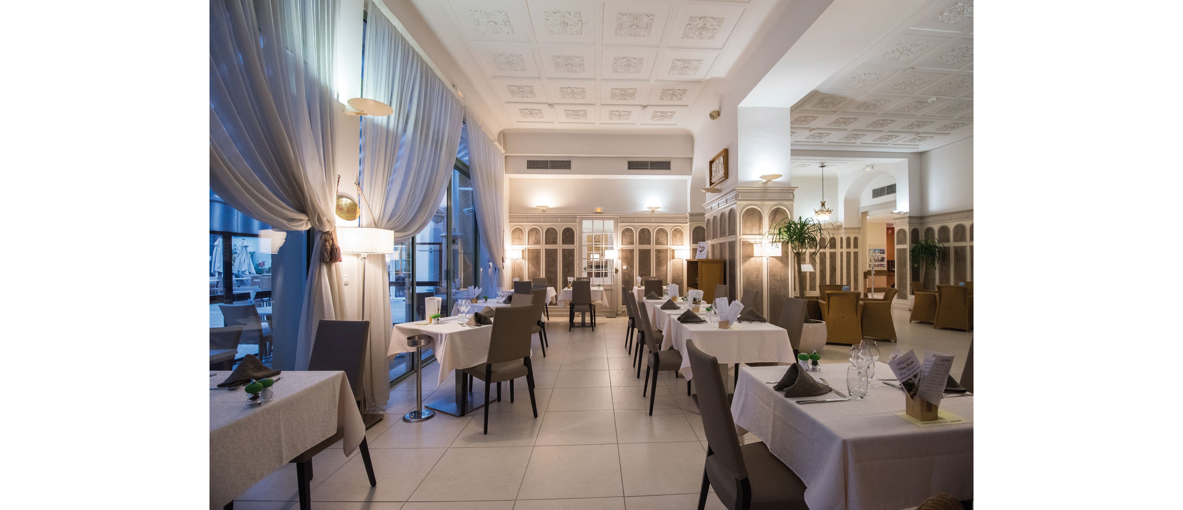 Upscale restaurant interior with neatly arranged tables and chairs is shown in soft evening lighting. White tablecloths, folded napkins, and glassware create a refined dining atmosphere. Large draped windows, decorative wall panels, and ceiling details add to the sophisticated design. The room appears quiet and empty, ready to welcome guests.