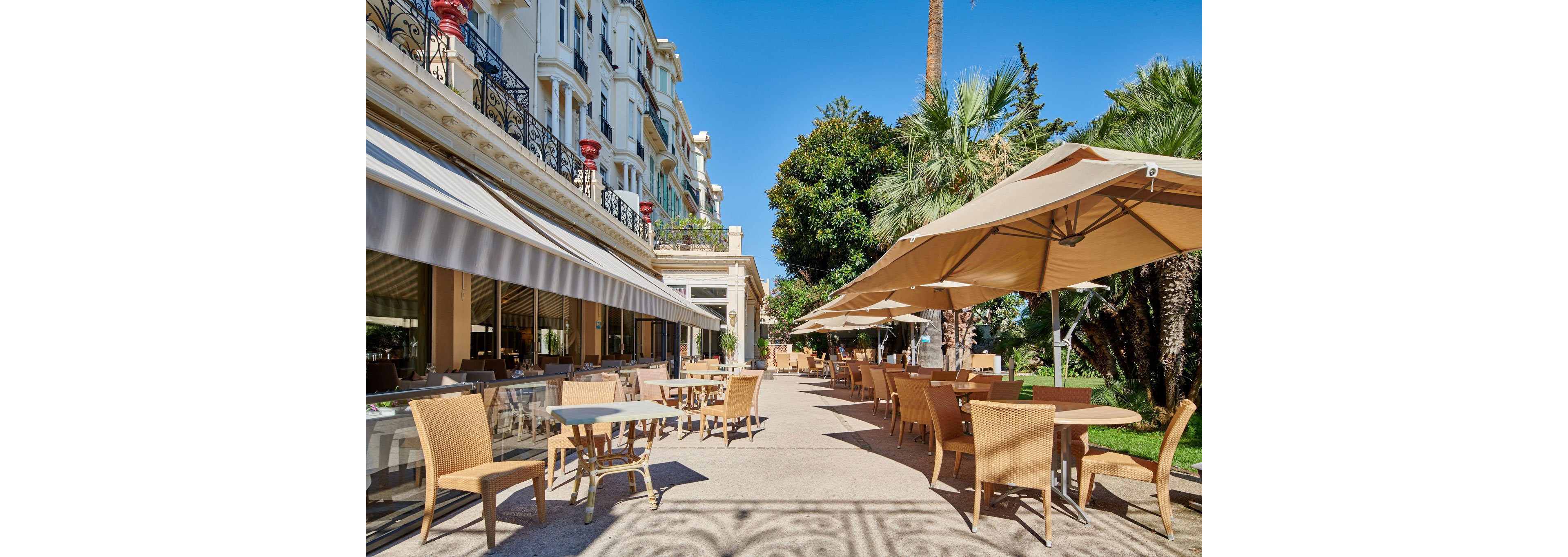 An elegant hotel terrace features numerous wicker chairs and tables arranged along a wide stone pathway. Large beige umbrellas provide shade beside lush green palm trees and manicured gardens. The multi-story building with balconies and striped awnings frames the scene under a clear blue sky. The setting appears calm and inviting, ideal for outdoor dining or relaxation.