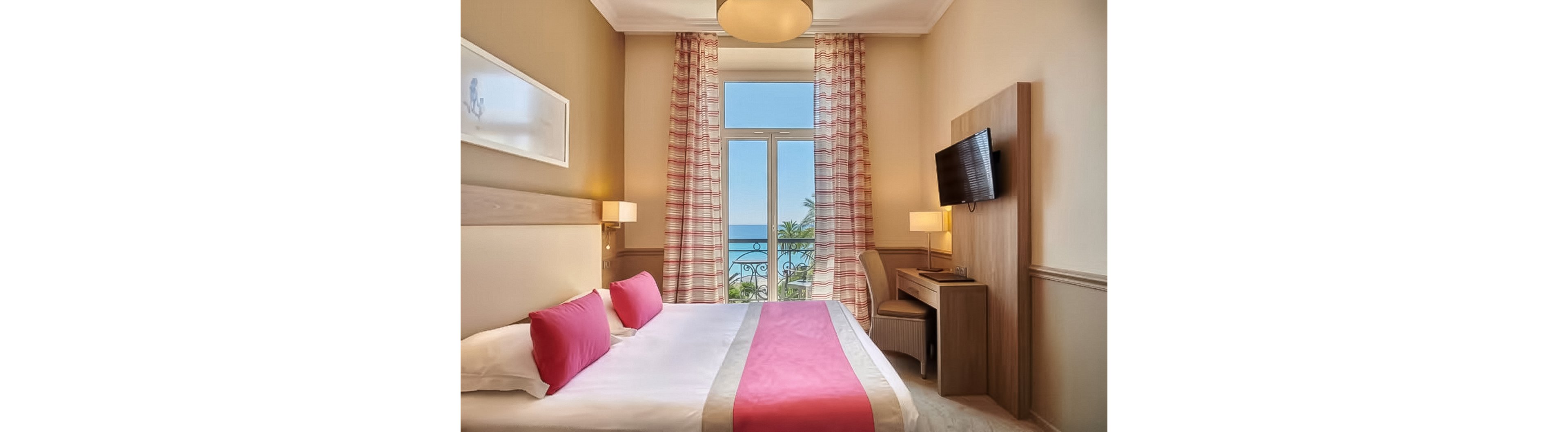 A neatly made hotel bedroom features a double bed with white linens and bright pink pillows and runner. Floor-to-ceiling windows with patterned curtains open to a balcony overlooking palm trees and a blue ocean. A wall-mounted television and compact wooden desk with chair sit to the right, while warm wall lamps provide soft lighting. The room has a clean, contemporary style with beige walls and a relaxed coastal atmosphere.