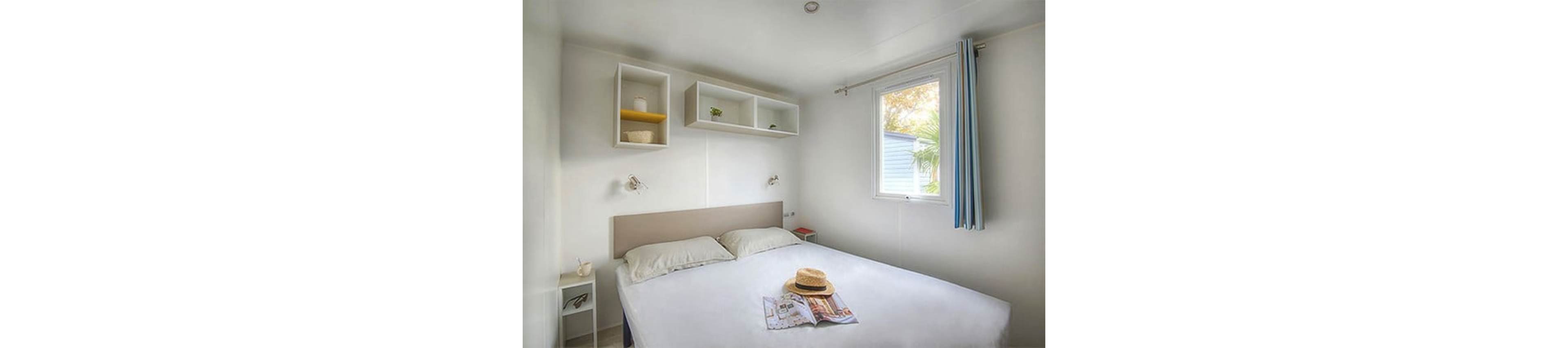 A clean and minimalist bedroom featuring a double bed with white linens and beige headboard. The room is well-lit with natural light streaming through a window adorned with blue-striped curtains. Decorative shelves and bedside tables add subtle accents, while a straw hat and magazine on the bed evoke a relaxed mood.