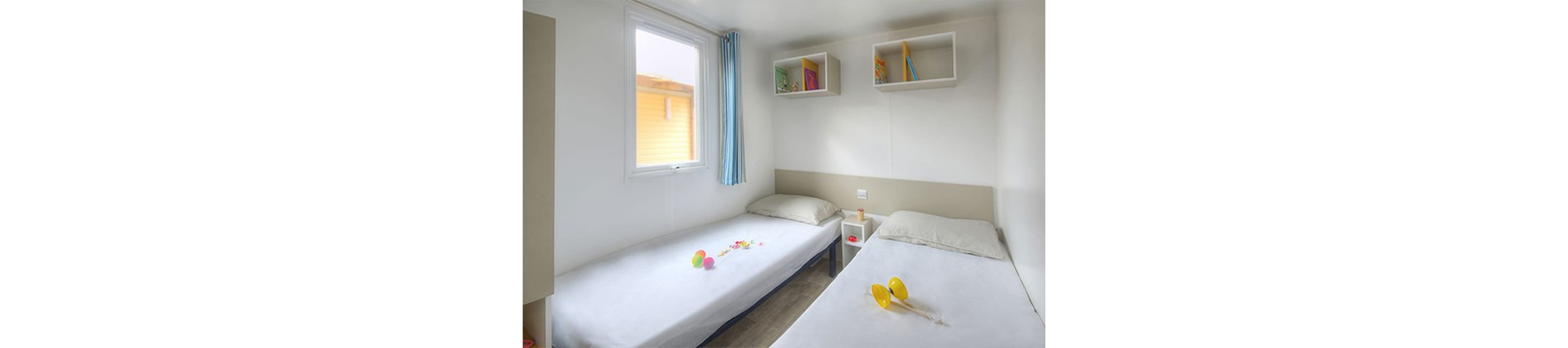 A compact twin bedroom featuring two single beds with neutral bedding. The room is brightly lit by a window with blue curtains, and colorful toys and books add playful accents. White shelves and a bedside table enhance the minimalist decor, while the yellow toy maracas stand out prominently.
