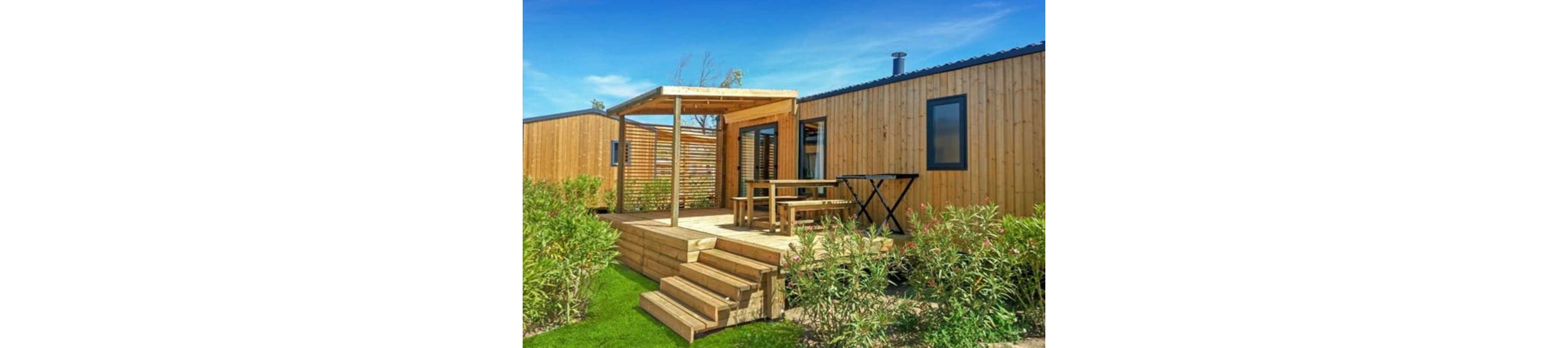 A contemporary wooden cabin featuring a spacious outdoor patio with a pergola. The setting includes a wooden table and bench, surrounded by lush greenery under a clear blue sky. The cabin has large windows and a sleek, minimalist design, ideal for relaxation or outdoor gatherings.