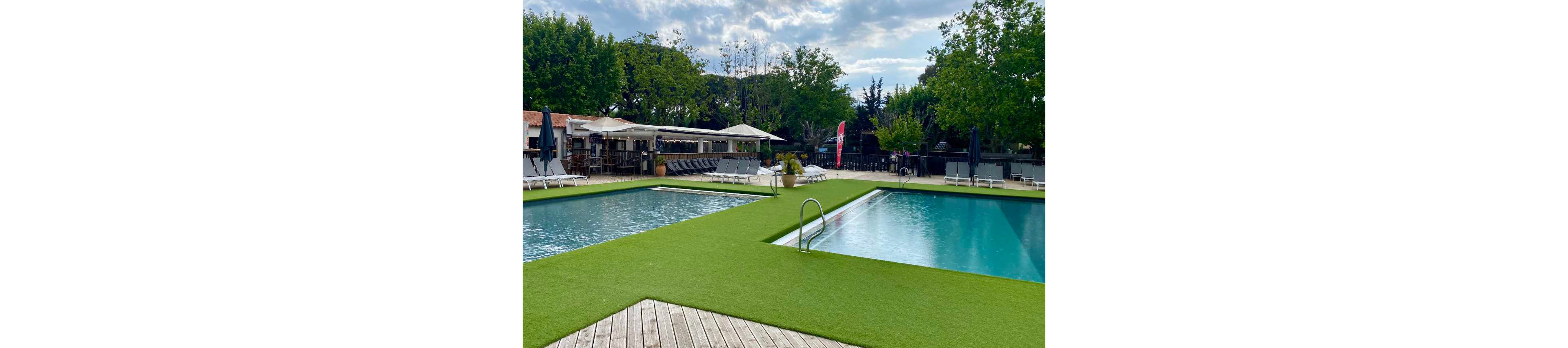 A serene outdoor pool area featuring two rectangular swimming pools surrounded by artificial grass and wooden decking. The setting includes lounge chairs, umbrellas, and a shaded bar area, creating a relaxing atmosphere. Tall trees and clear skies enhance the tranquil environment.