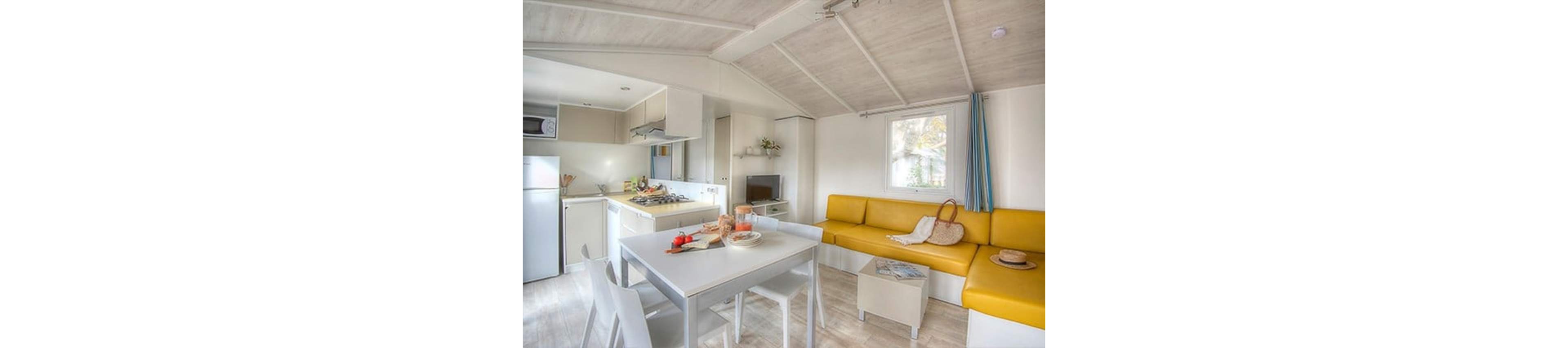 A cozy and modern kitchen and living area featuring a yellow sofa, white dining table, and compact kitchen appliances. The space is well-lit with natural light streaming through the windows. Minimalist decor includes a small TV, a microwave, and a fruit bowl on the table. The setting exudes a clean and inviting atmosphere, perfect for relaxation or casual dining.