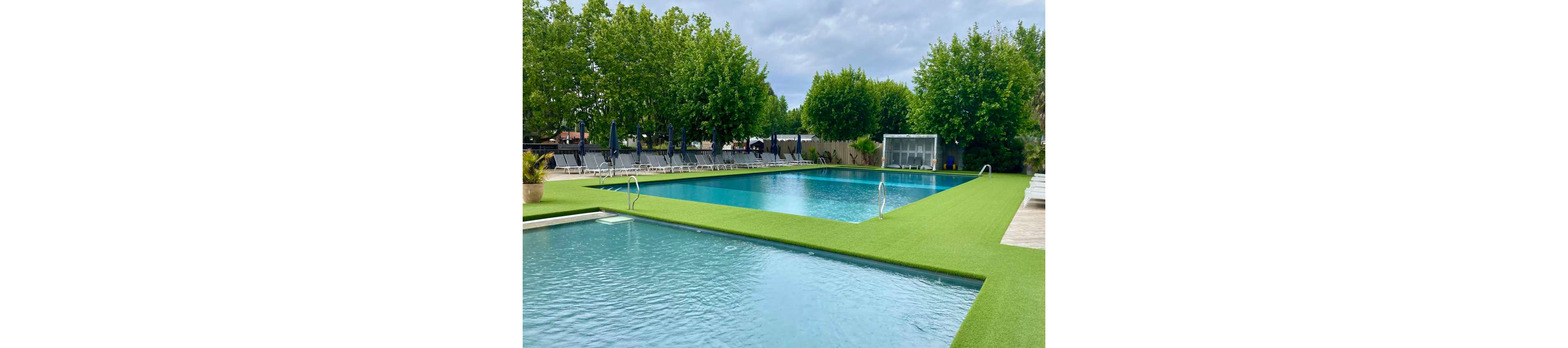 A spacious outdoor pool area featuring two pools surrounded by vibrant green artificial grass. The setting includes lounge chairs and umbrellas, ideal for relaxation. Tall trees and cloudy skies create a serene atmosphere, perfect for leisure activities.