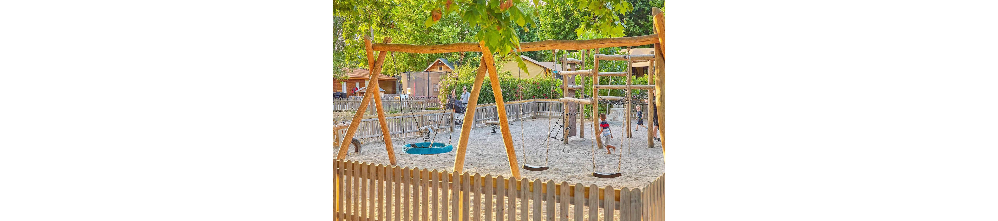 A vibrant outdoor playground featuring wooden swings and climbing structures surrounded by a wooden fence. The setting is sunny with lush green trees and a sandy ground. Children are actively playing, and adults are seen in the background. The scene conveys a lively and family-friendly atmosphere.