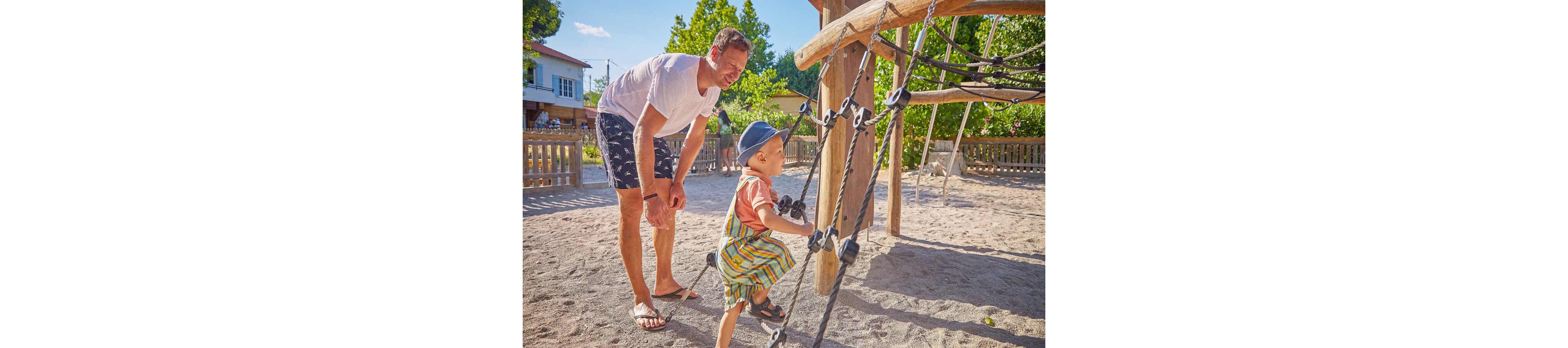 A young child wearing a striped outfit and hat climbs a rope structure at an outdoor playground. An adult stands nearby, offering support. The setting is sunny with trees, a wooden fence, and residential houses in the background.