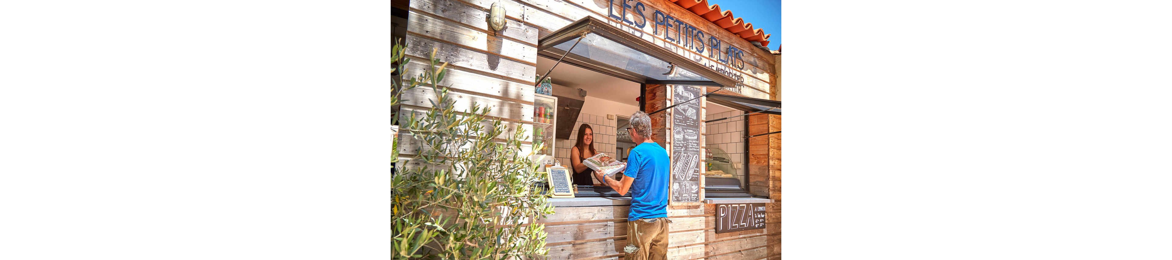 A man in a blue shirt is purchasing pizza at an outdoor wooden food stand labeled 'Les Petits Plats'. The setting is bright and sunny, with olive plants visible in the foreground. The stand features rustic wooden panels and a menu board, creating a casual and inviting atmosphere.