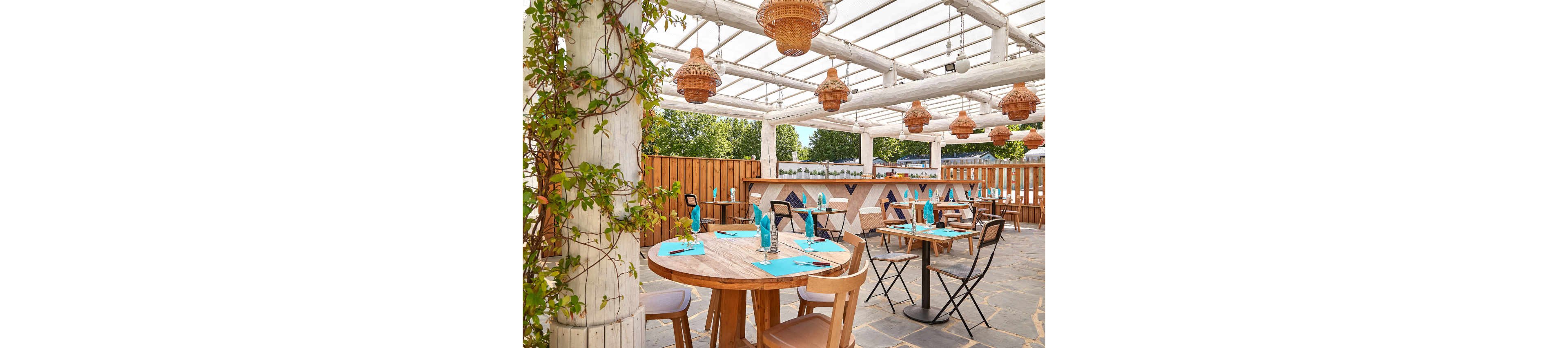 A spacious outdoor dining area featuring wooden tables and chairs under a white pergola. The setting includes hanging wicker lamps and turquoise table settings, creating a vibrant and inviting atmosphere. Greenery and natural light enhance the rustic charm of the space.