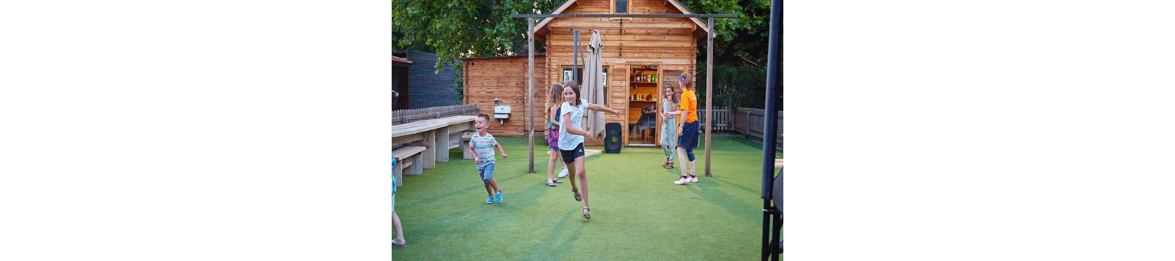 A group of children and teenagers are seen playing on a grassy outdoor area near a wooden cabin. The setting includes picnic-style benches and a small structure resembling a shed or cabin. The mood is lively and cheerful, with kids running and interacting in a casual environment. The scene is illuminated by natural light, emphasizing the warm and inviting atmosphere.