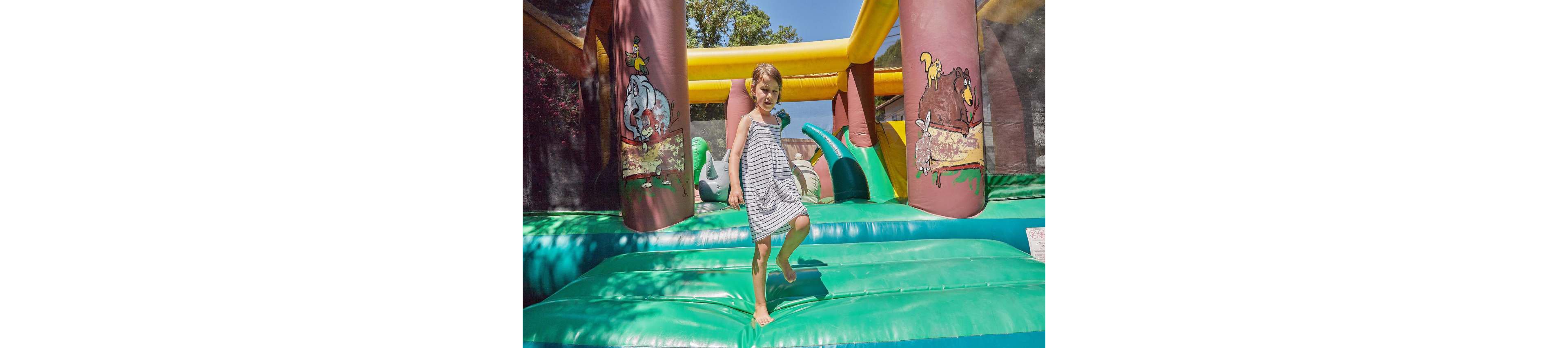 A young child is seen enjoying an inflatable bounce house in an outdoor setting. The bounce house features colorful cartoon animal designs on its pillars, including an elephant and a bear. The sunny day and vibrant colors create a playful and cheerful atmosphere.