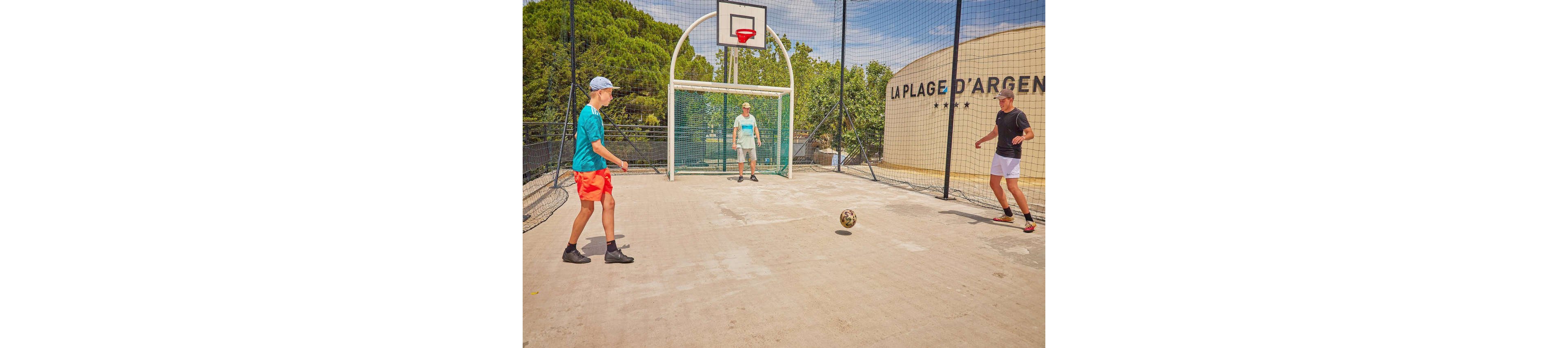 Three young men play soccer on a concrete court with a basketball hoop in the background. The setting is outdoors, surrounded by greenery and a visible building labeled 'La Plage d'Argent'. The players are dressed in casual sportswear, engaging in a lively activity under clear skies.