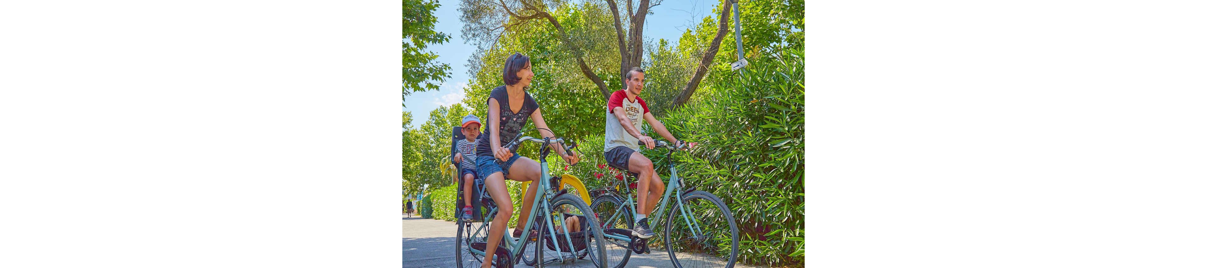 A family enjoys a leisurely bike ride on a sunny day in a lush, green outdoor setting. The adults ride bicycles while a child is seated in a child bike seat. The path is surrounded by trees and bushes, creating a serene and natural environment. The scene captures an active and healthy lifestyle in a vibrant, sunny atmosphere.