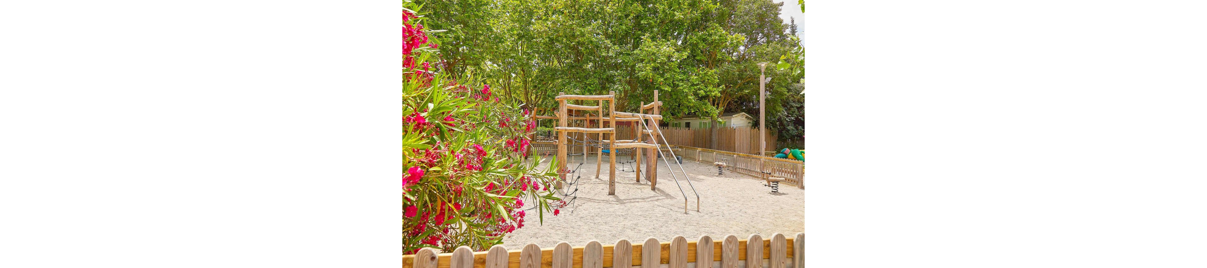 A wooden playground structure with a slide is set in a sandy area surrounded by a wooden fence. The scene is bright and sunny, with lush green trees and vibrant pink flowers adding a natural touch. The setting appears to be a quiet outdoor park or play area, ideal for children.