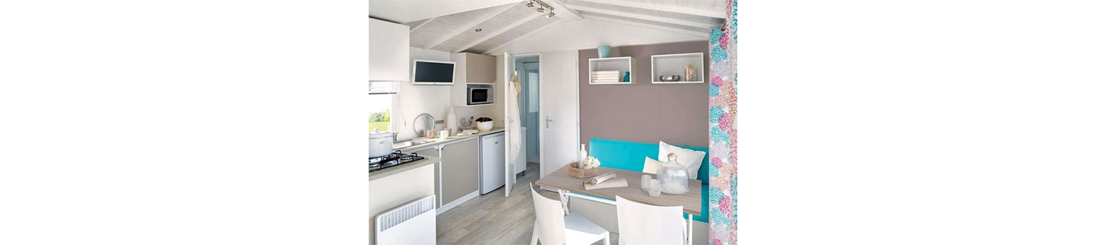 A bright and modern compact kitchen and dining area featuring neutral tones and pops of turquoise. The setting includes a small stove, microwave, and refrigerator, with a dining table set for two. The space is well-lit with natural light and decorated with colorful curtains and minimalist shelving.