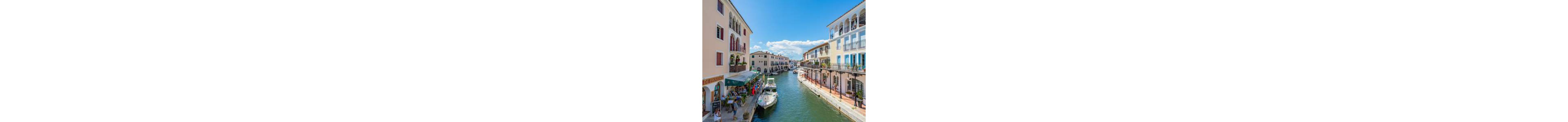 A picturesque canal is lined with pastel-colored buildings and outdoor cafes on a bright sunny day. Small boats are moored along the waterway, with one boat in the foreground near a bustling terrace. People sit and walk along the waterfront, enjoying the pleasant weather and relaxed atmosphere. Balconies, awnings, and clear blue skies enhance the Mediterranean-style charm of the scene.