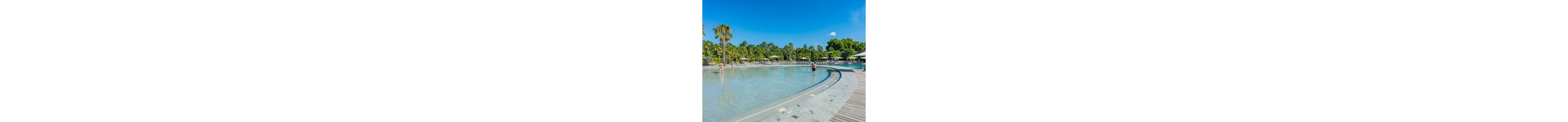 A large shallow resort swimming pool curves alongside a wooden deck under a clear blue sky. Several people relax and wade in the water while others rest on loungers shaded by white umbrellas. Tall palm trees and lush green foliage surround the pool, creating a tropical atmosphere. The bright sunlight and clear weather emphasize a calm, leisurely vacation setting.