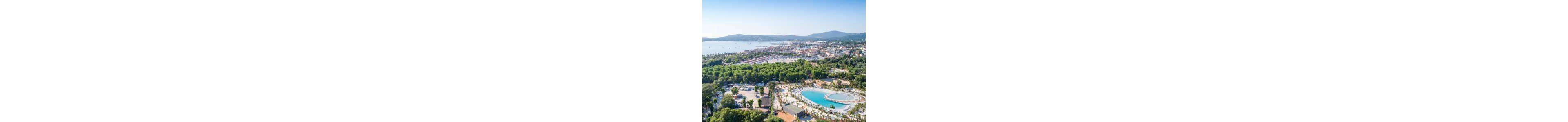 An aerial view showcases a coastal resort town with a large lagoon-style swimming pool in the foreground. The resort is surrounded by lush green trees and low-rise buildings. Beyond the resort, a marina and calm bay with anchored boats stretch toward the horizon. Rolling hills and distant mountains frame the background under a clear blue sky, creating a serene holiday atmosphere.