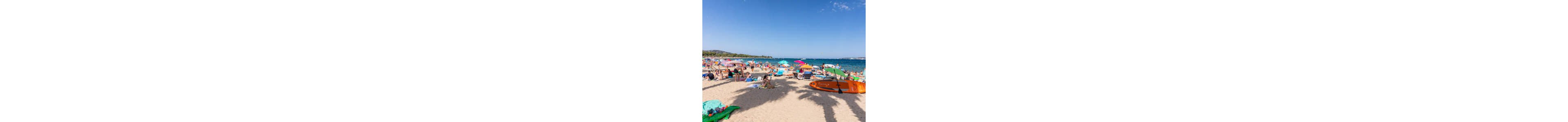 A busy sandy beach is filled with sunbathers, colorful umbrellas, and beach towels under a clear blue sky. People of various ages relax, swim, and play near the shoreline as waves roll in. An orange kayak and other beach gear are visible in the foreground, adding to the active holiday atmosphere. The distant coastline and calm sea create a vibrant summer vacation scene.