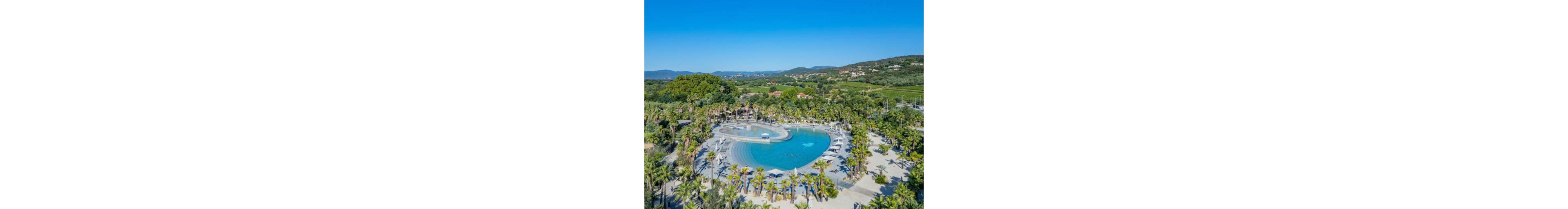 An aerial view showcases a large, lagoon-style swimming pool surrounded by palm trees. Sun loungers and umbrellas line the curved pool edges, suggesting a relaxing vacation setting. The pool sits within a lush green landscape with hills and scattered buildings in the background. Clear blue skies and bright sunlight create a warm, summery atmosphere.