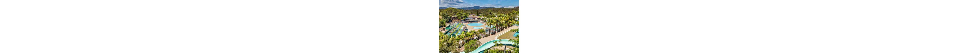 An expansive resort pool complex is surrounded by rows of sun loungers and umbrellas. Multiple circular and oval swimming pools sit amid palm trees and lush greenery. A curved waterslide winds through the foreground, leading toward the pool area. In the background, forested hills and distant mountains create a scenic natural backdrop under a bright, clear sky.