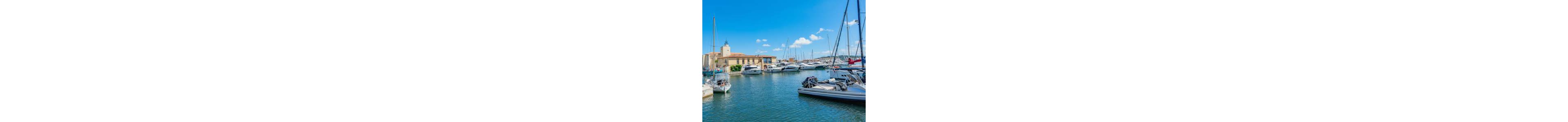 A bright marina scene shows several yachts and sailboats moored in calm blue water. A beige waterfront building with a small tower stands along the quay under a clear sky. Masts and rigging create vertical lines against the horizon, with more boats visible in the background. The overall atmosphere is peaceful and summery, suggesting a leisurely coastal destination.