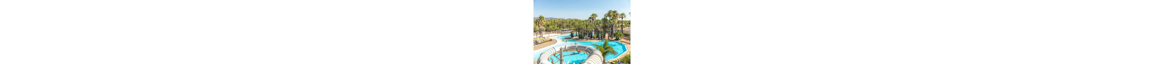 A large outdoor swimming pool winds through a sunny resort surrounded by tall palm trees. A curved waterslide empties into the bright blue water, adding a sense of fun and activity. Sun loungers line the poolside where guests relax under the clear sky. Low white buildings and distant hills frame the tropical holiday setting.