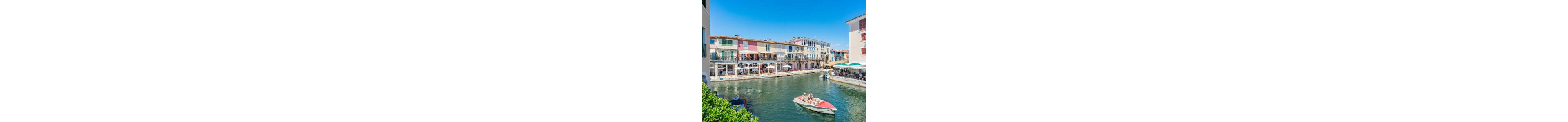 A small motorboat with two people glides along a calm canal in front of colorful waterfront buildings. The pastel facades feature balconies, awnings, and large windows overlooking the water. Outdoor restaurant terraces line the quay, with diners seated under umbrellas. The bright blue sky and vivid sunlight create a cheerful, summery atmosphere in this picturesque canal-side scene.